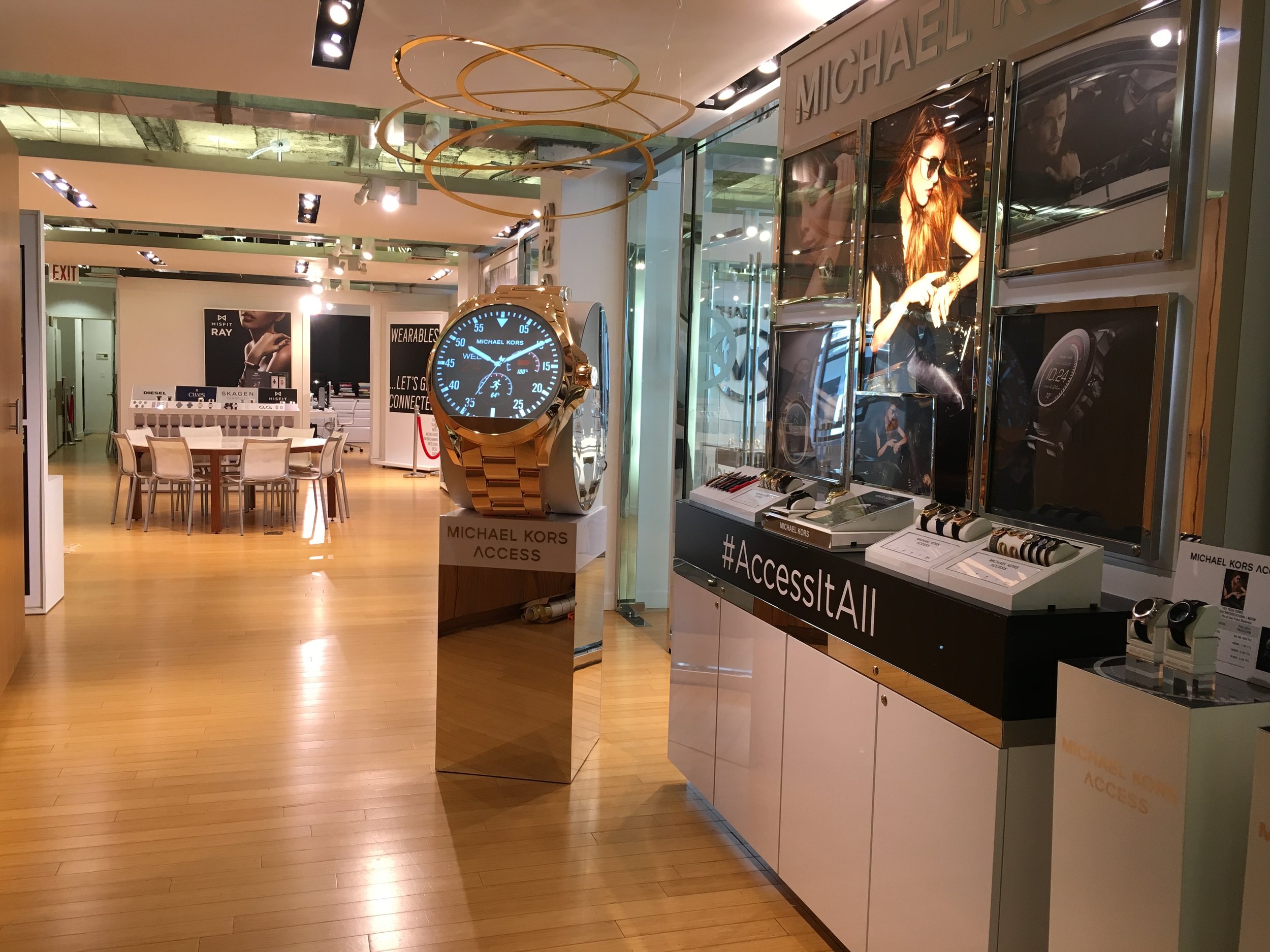 Interior of a retail store selling Michael Kors accessories and watches, with display cases, promotional posters, and a large golden watch on a mirrored stand.