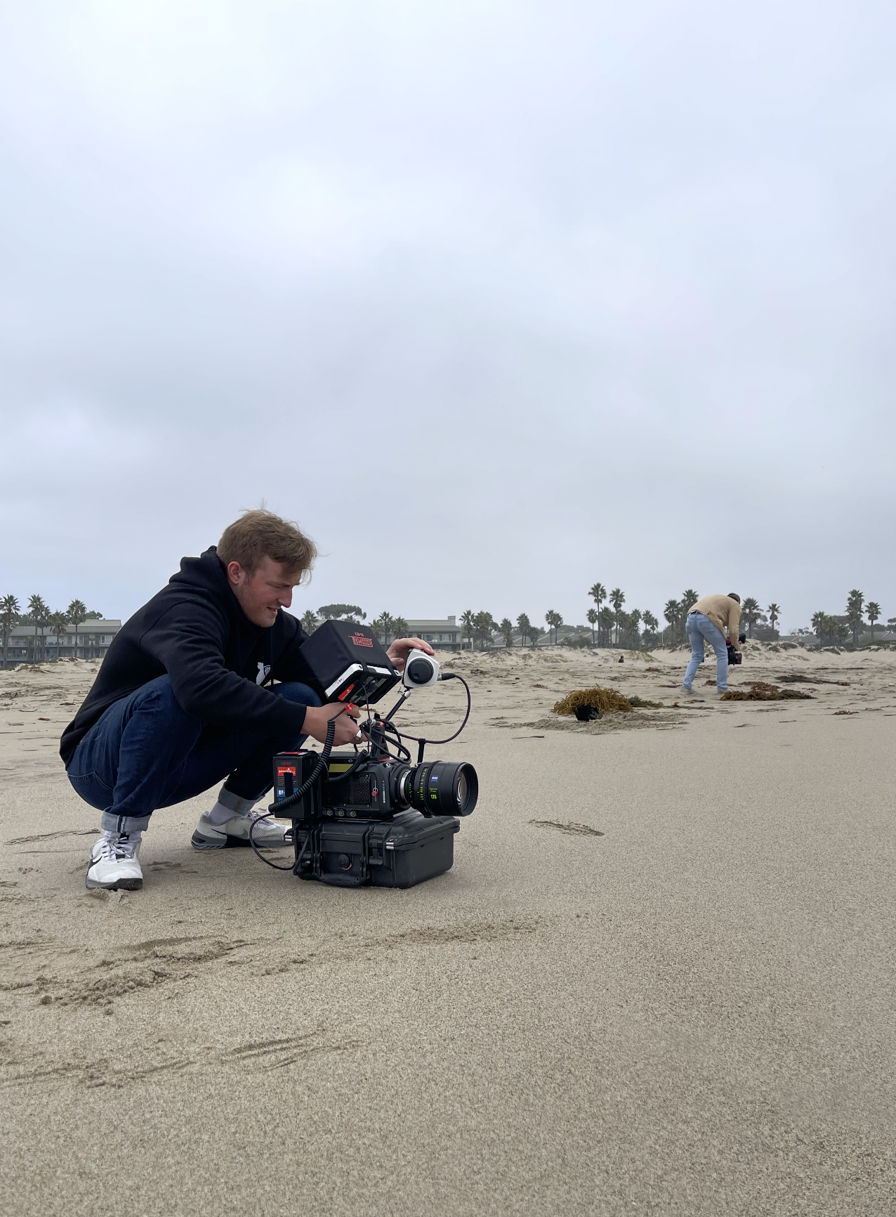 A person crouching on a sandy beach with a professional video camera, while another person in the background is examining the sand, under a cloudy sky.