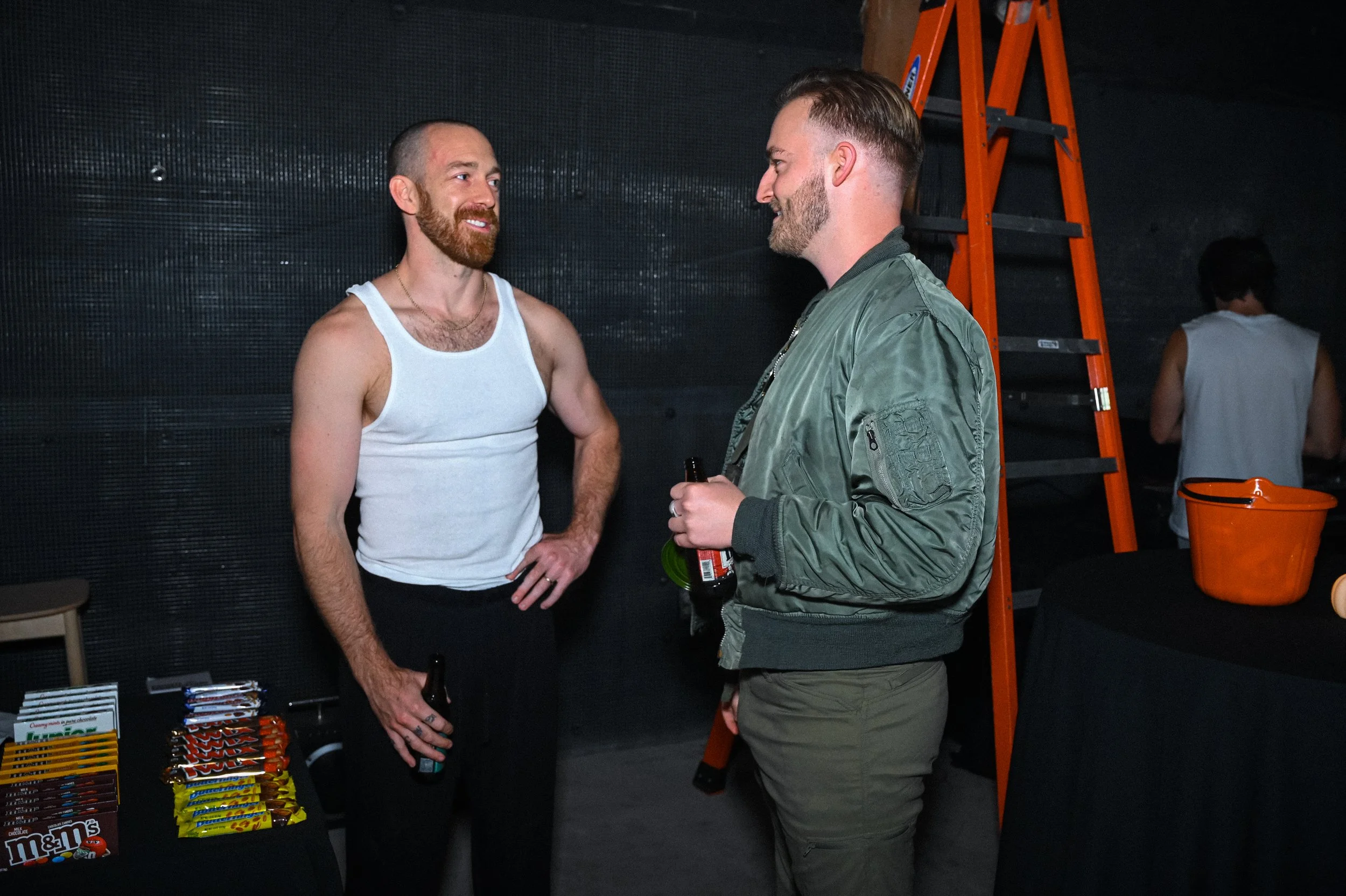 Two men having a conversation at a social event, standing near a table with candy and snacks, in a room with dark walls and a ladder in the background.