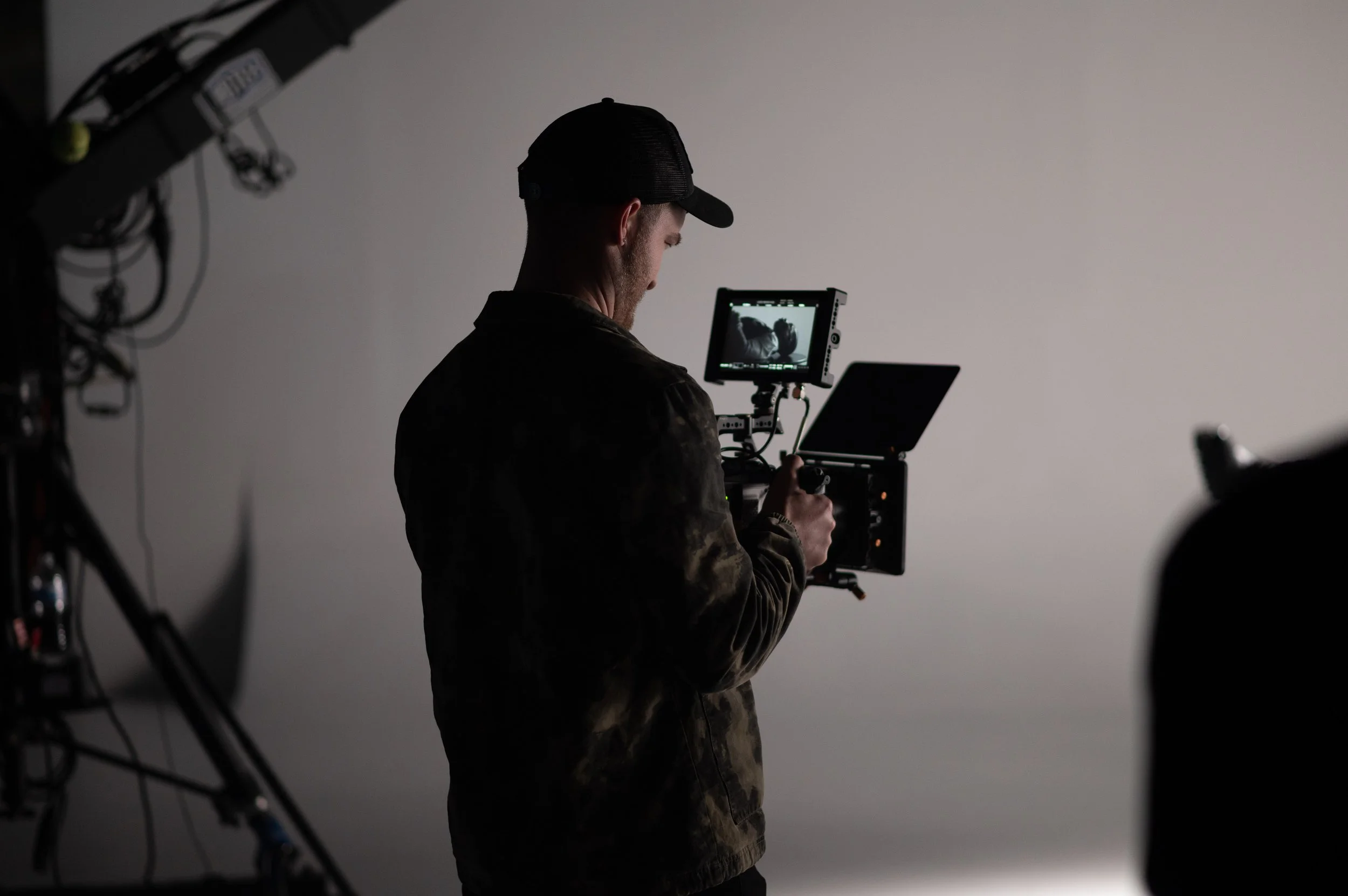 Person in a dark cap and camouflage jacket operating a professional video camera on a light grey studio background.