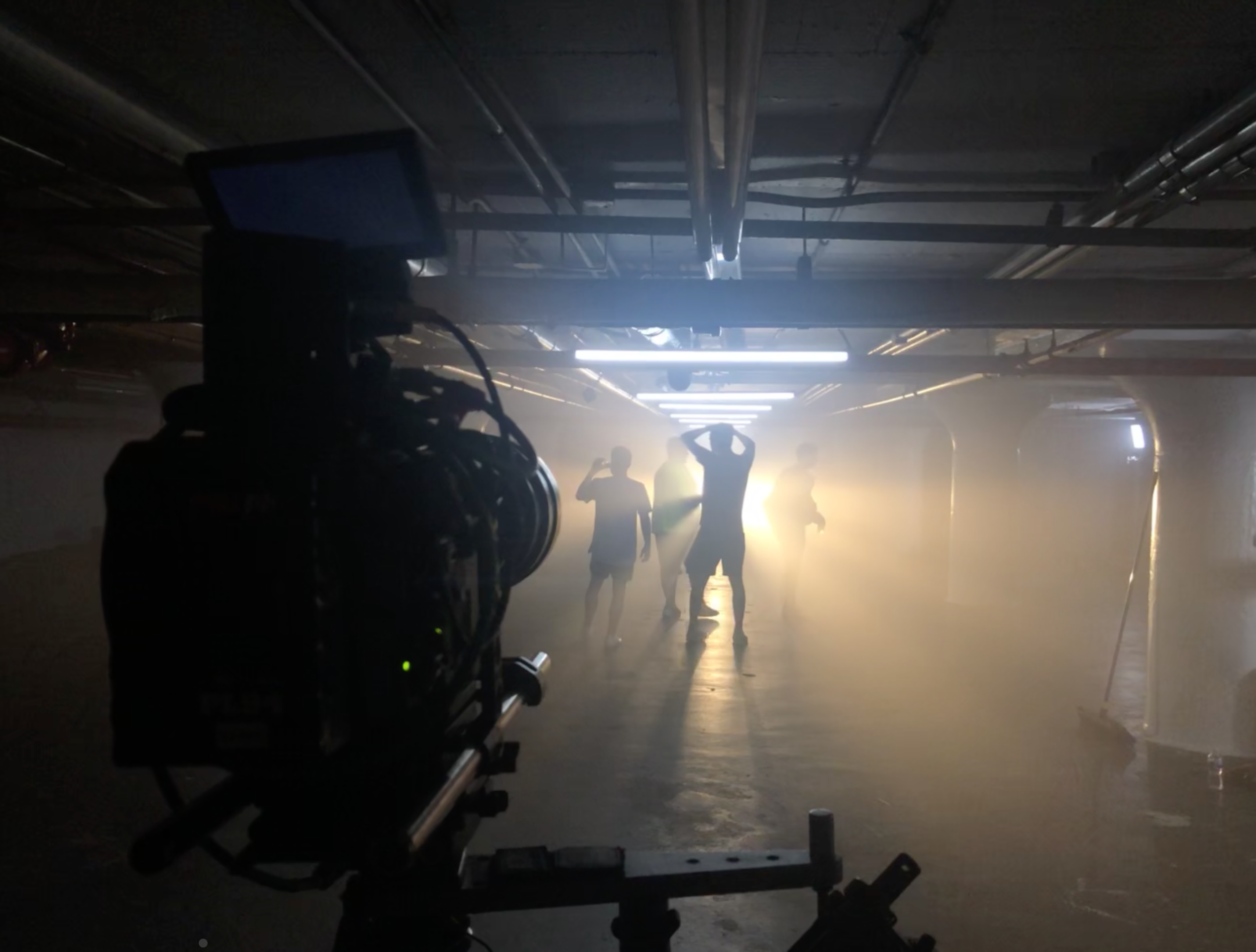 Film crew setting up shot in a dimly lit underground parking garage with sunlight filtering in, silhouetting people, and a camera on a dolly in foreground.