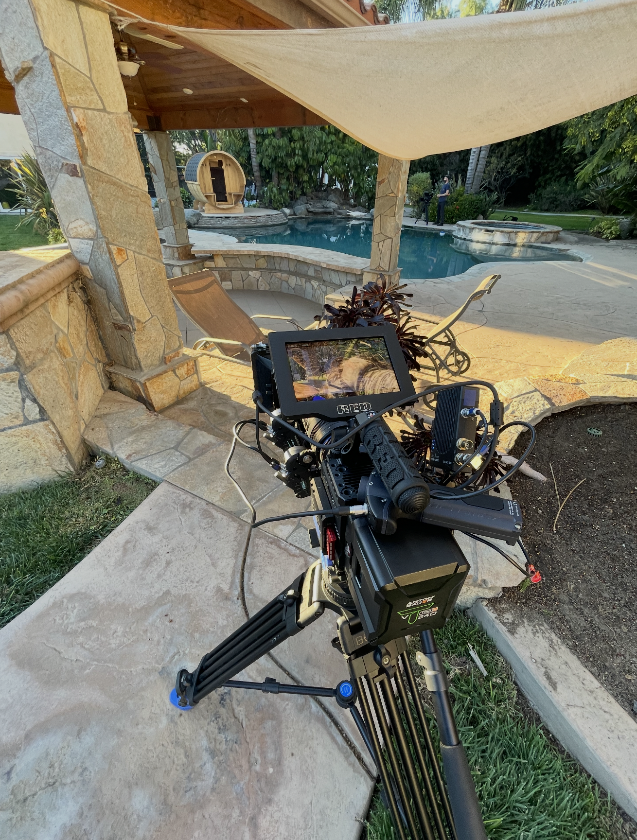 A professional video camera set up on a tripod capturing footage of an outdoor backyard pool area with lounge chairs, stone structure, a wooden shaded pavilion, and lush green foliage in the background.
