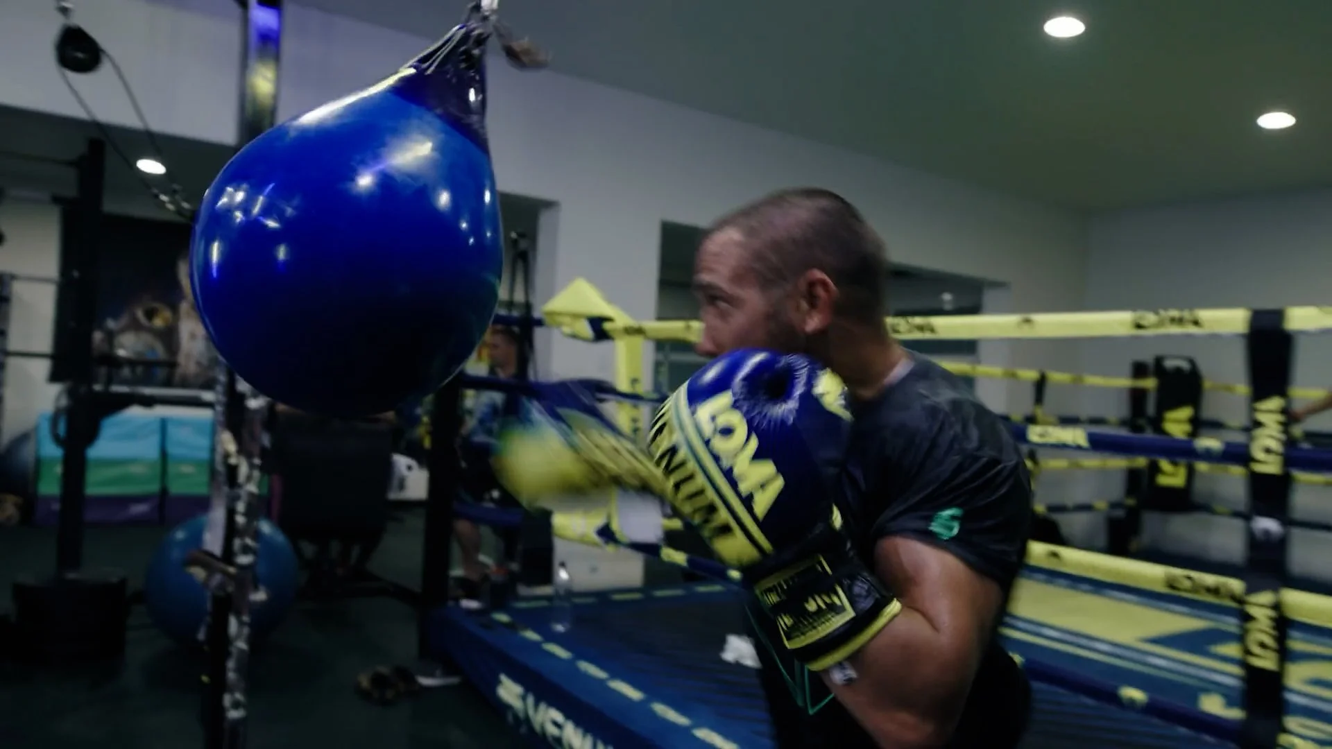 A man practicing boxing with a blue punching bag in a gym, wearing boxing gloves and black athletic clothing, with boxing ring and equipment in the background.
