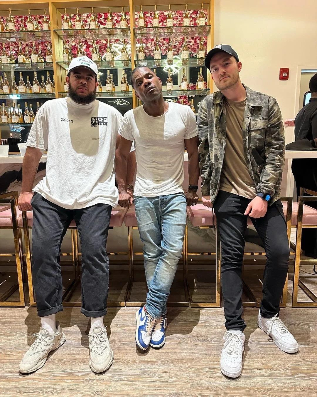 Three young men standing in front of a bar with pink floral wallpaper and bottles of champagne on display. They are dressed casually and wearing sneakers.