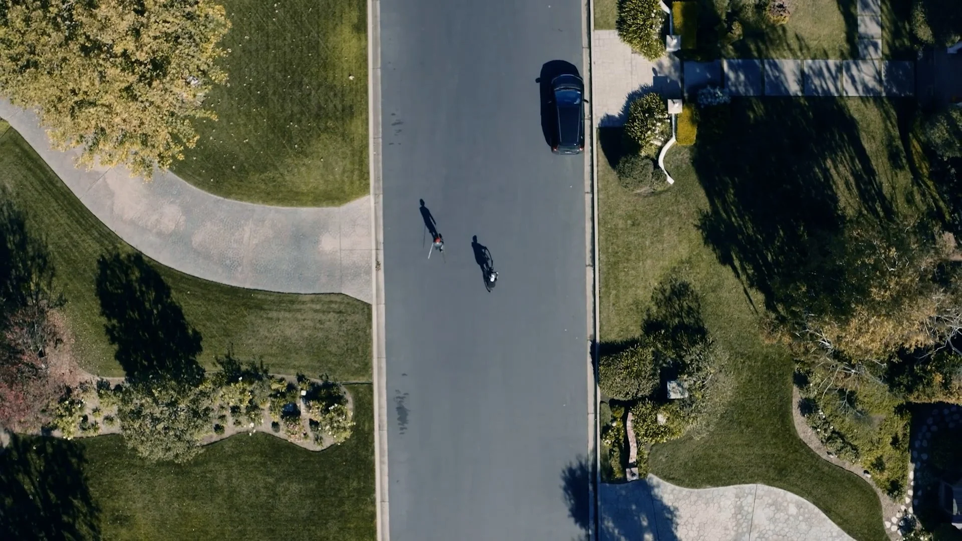 An aerial view of a residential neighborhood showing a driveway with two people riding bicycles and a parked car, surrounded by green lawns and landscaped gardens.