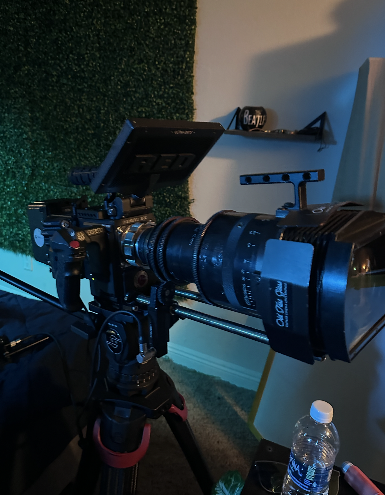 A professional camera mounted on a tripod, with a large telephoto lens, in a room with a green artificial grass wall and shelves in the background.
