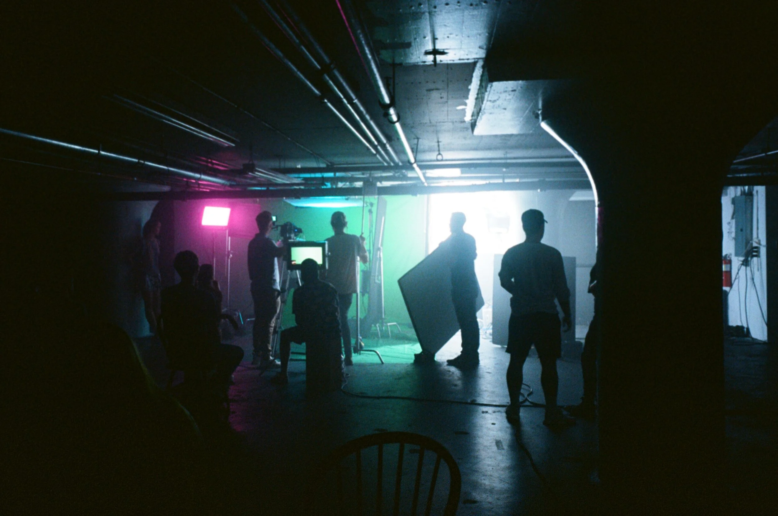 Behind-the-scenes of a film or photo production with crew members working in a dimly lit space, illuminated by colorful studio lights and a large green screen.