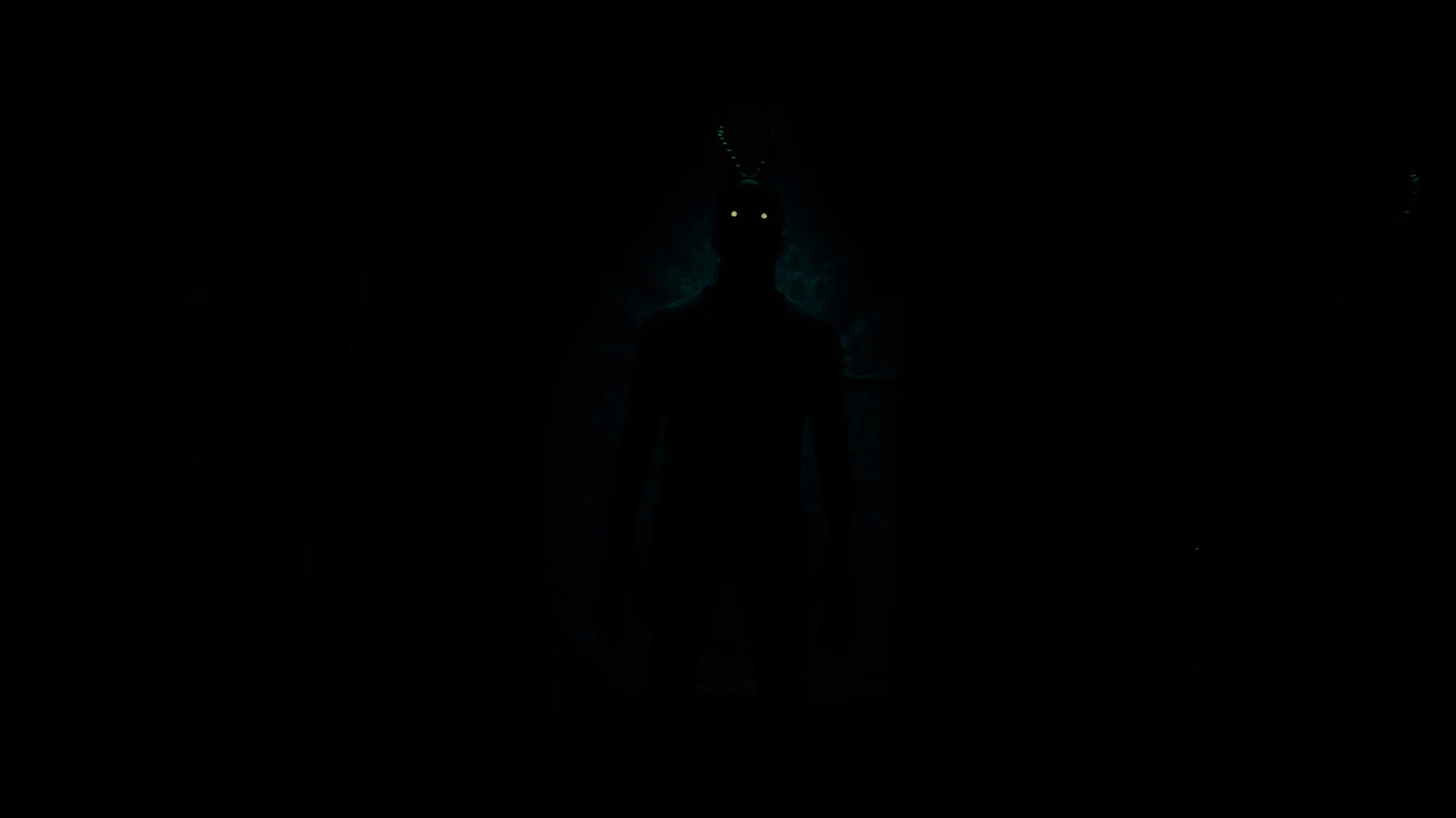 A dark, mysterious figure standing with glowing eyes in a shadowy environment, with a faint blue-green glow behind them and what appears to be antlers or horns on their head.