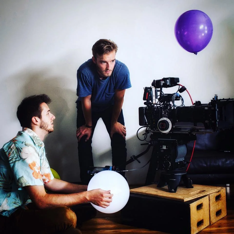 Two young men working on a film set. One sitting with a balloon in his hand, the other leaning over and looking at a large professional camera equipment. A purple balloon is floating in the air.
