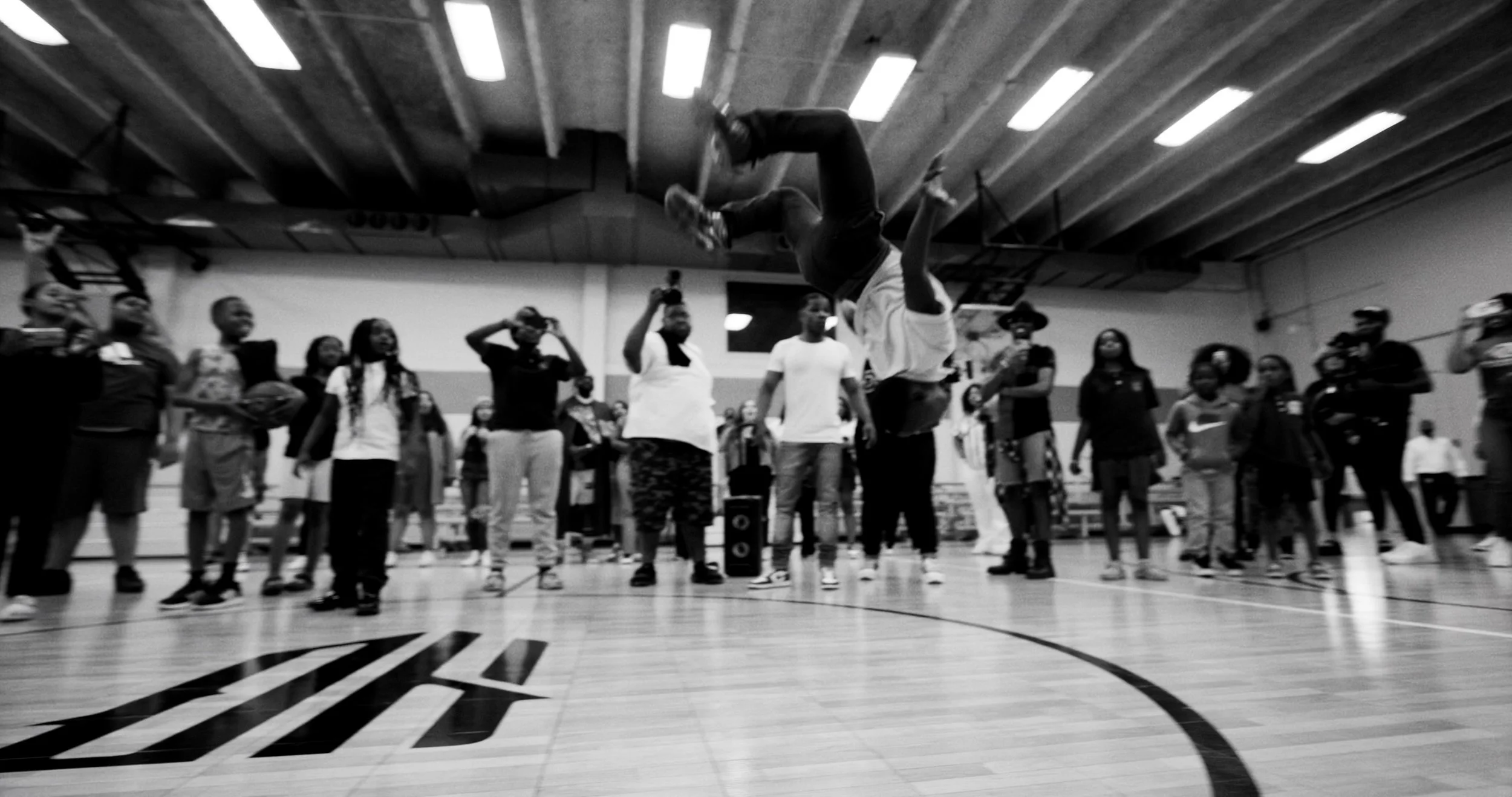 A basketball player performing a flip mid-air during a demonstration or event in a gymnasium filled with children and spectators watching.