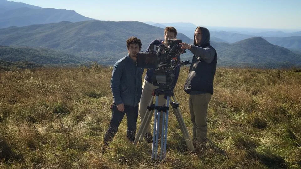 Three men setting up a camera on a tripod in a grassy field with mountain ranges in the background, under a clear sky.