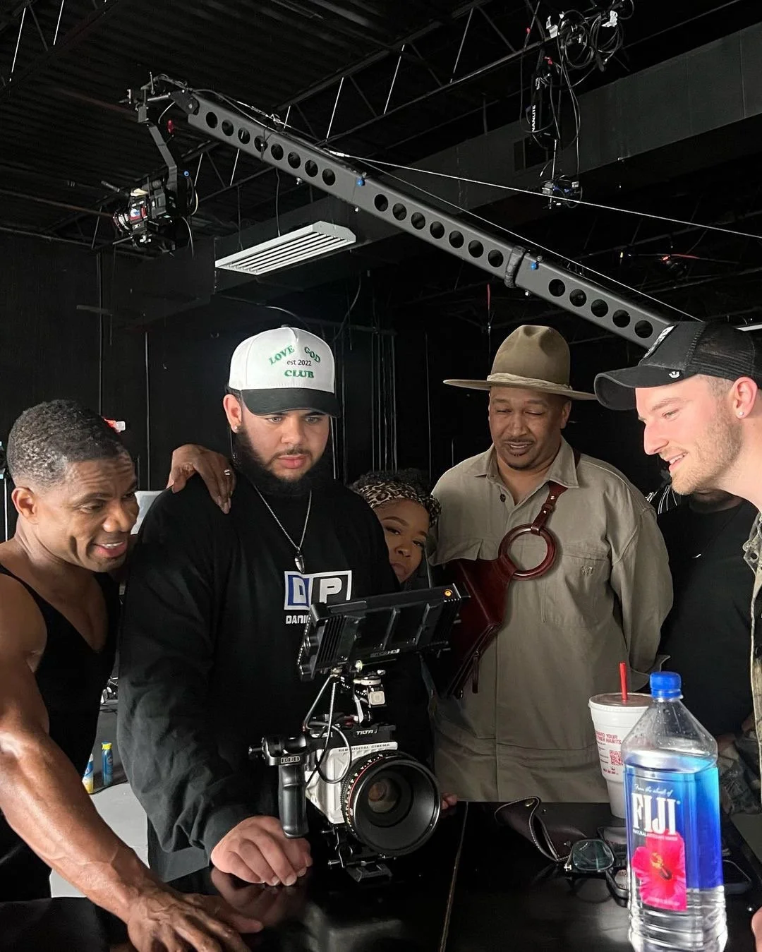 Group of five people gathered around a camera on a film set, with lighting and equipment overhead, and drinks on the table.