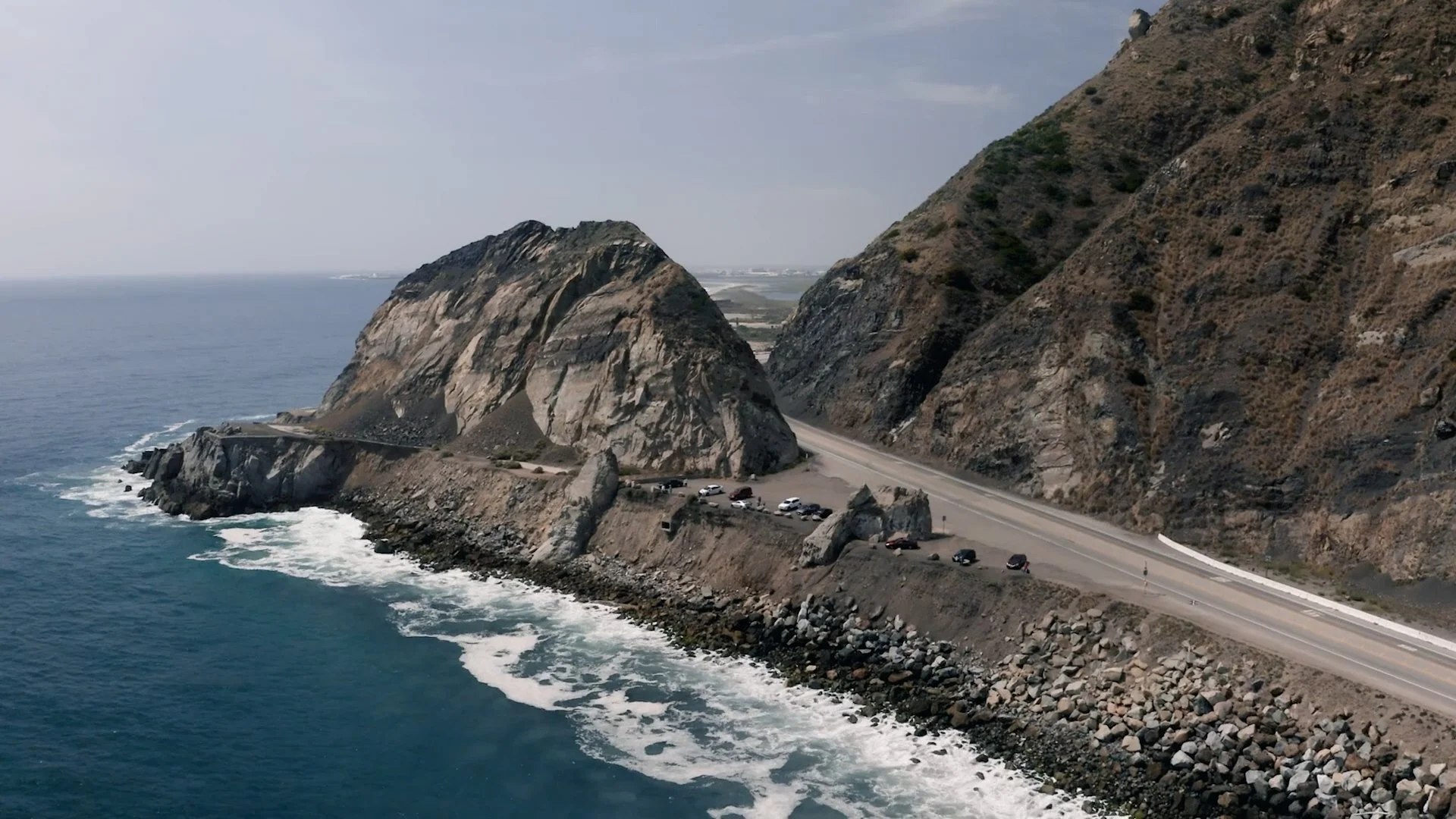 A coastal highway runs along a rocky cliffside with large rocks and dry vegetation; the ocean is visible on the left, with waves crashing against the rocks.