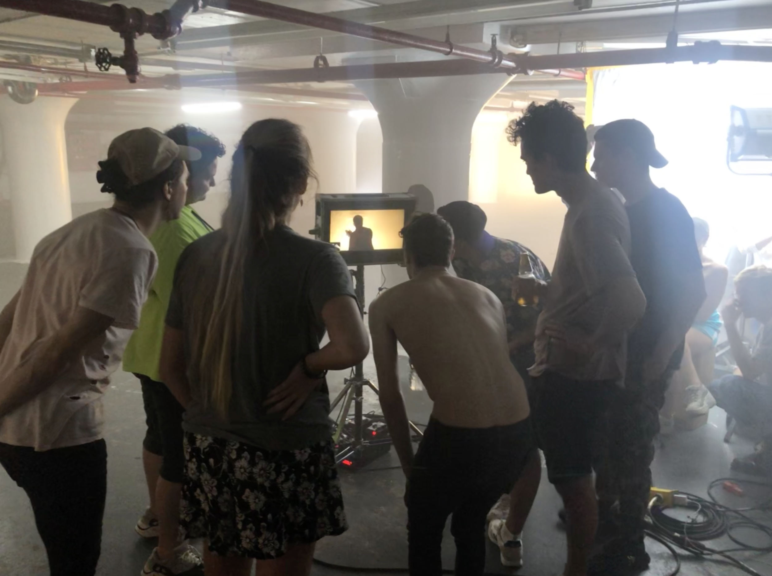 Film crew gathered around a monitor on set, with a silhouette of a person on the screen, in a foggy or smoky indoor location.