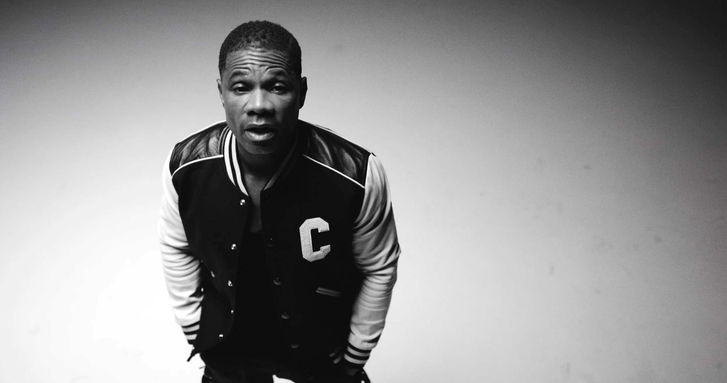 Black and white photo of a young man with short hair, wearing a varsity jacket with a large letter 'C' on it, standing in a sparsely decorated room with a plain background.