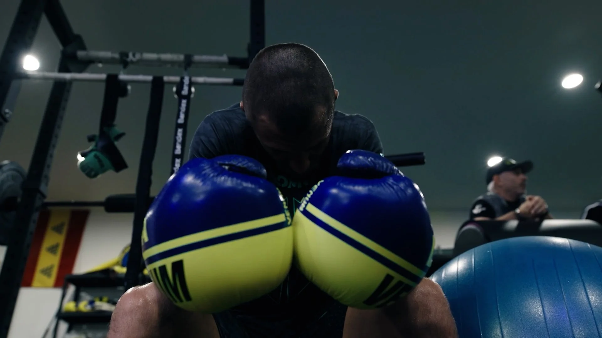 Boxer in a gym with blue and yellow gloves resting on his knees, head bowed, and a ball in front of him.