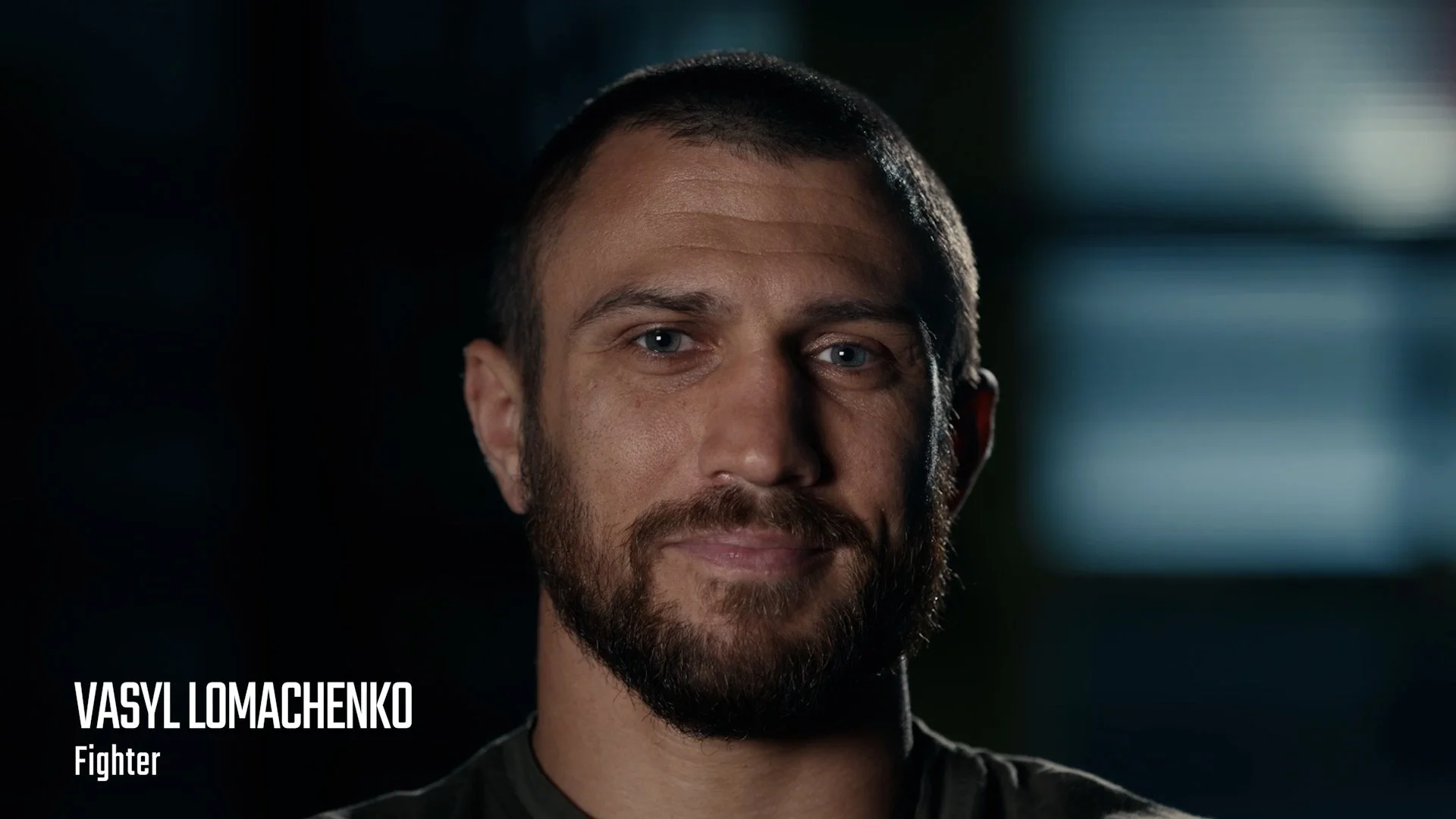 A man with short brown hair and a beard, wearing a dark shirt, looking at the camera in a dimly lit environment. Text overlay states: 'Vasyl Lomachenko, Fighter'.