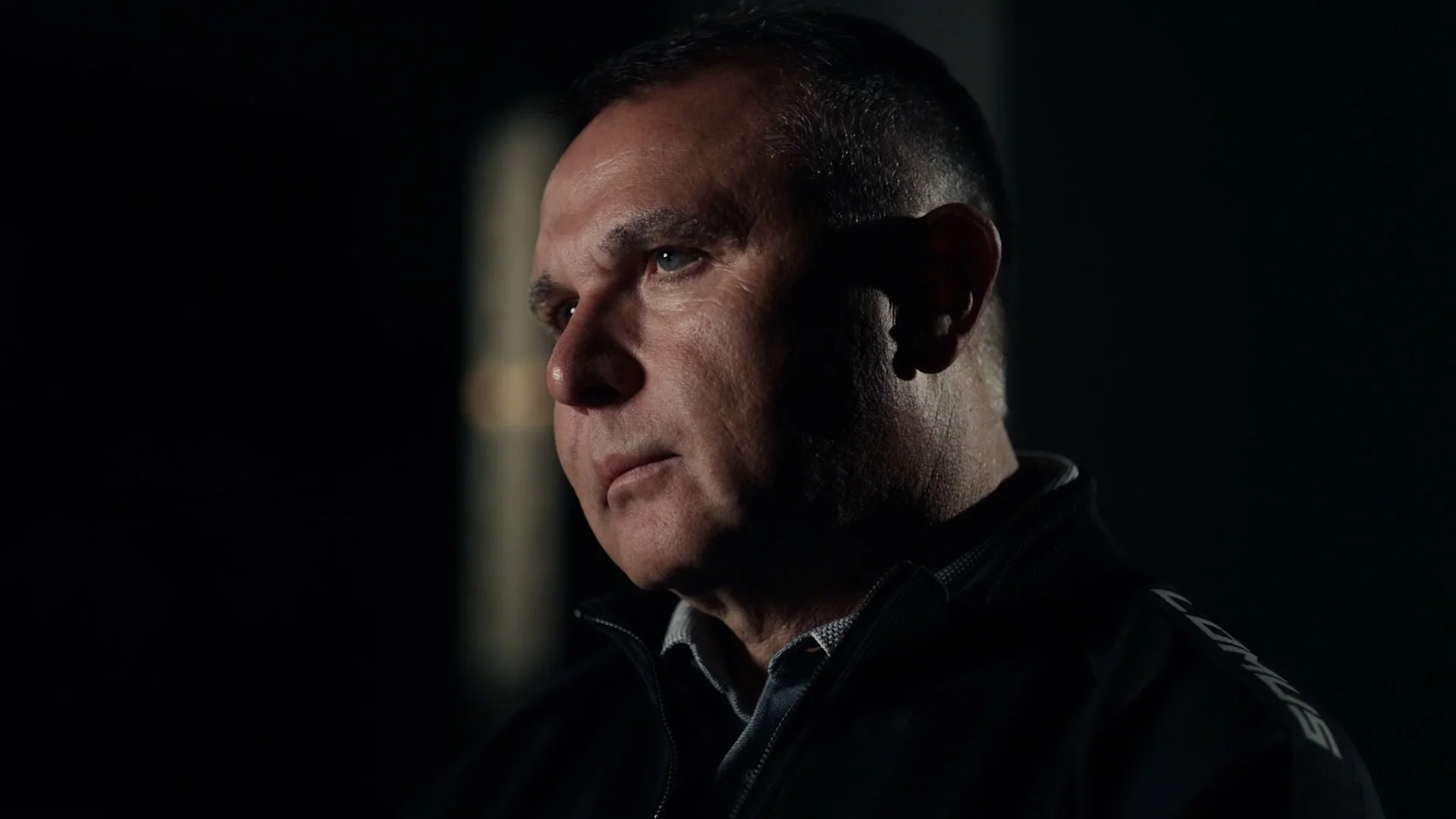 Close-up of a man with short dark hair, looking to the side with a serious expression, in front of a dark background with a blurred light source.