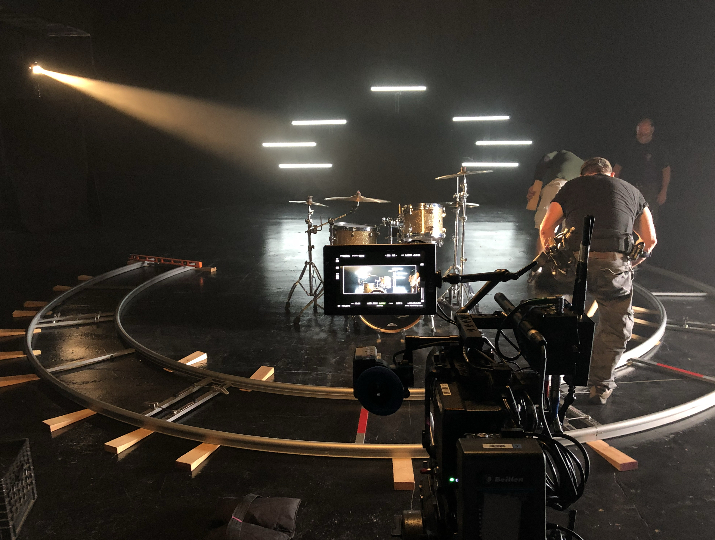 A musical drum set positioned on a circular track, with crew members adjusting equipment around it, and a camera capturing the scene, in a dark studio setting with bright light beams in the background.