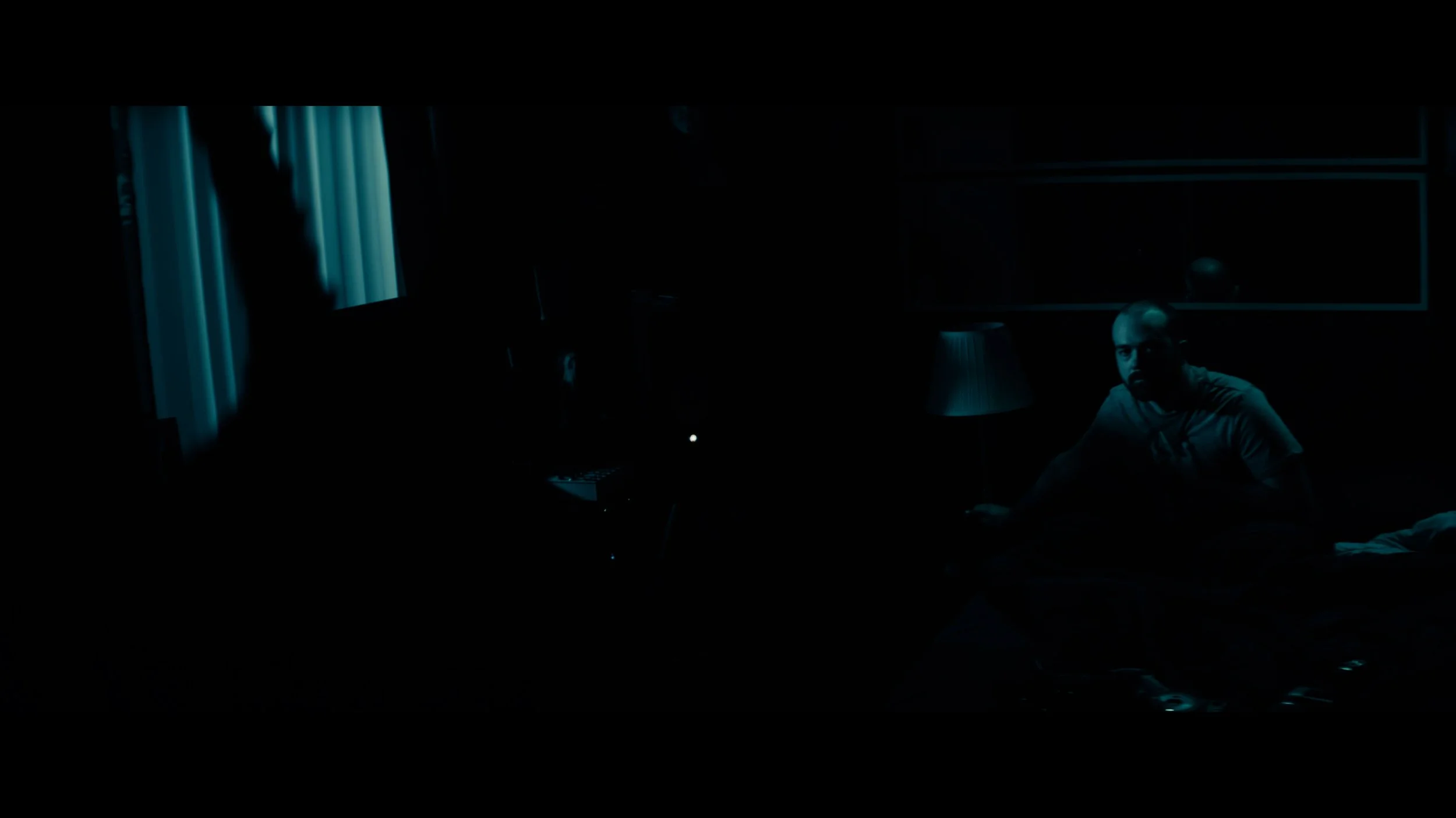 A man sitting on a bed in a dark room lit by blue light, looking towards the camera.