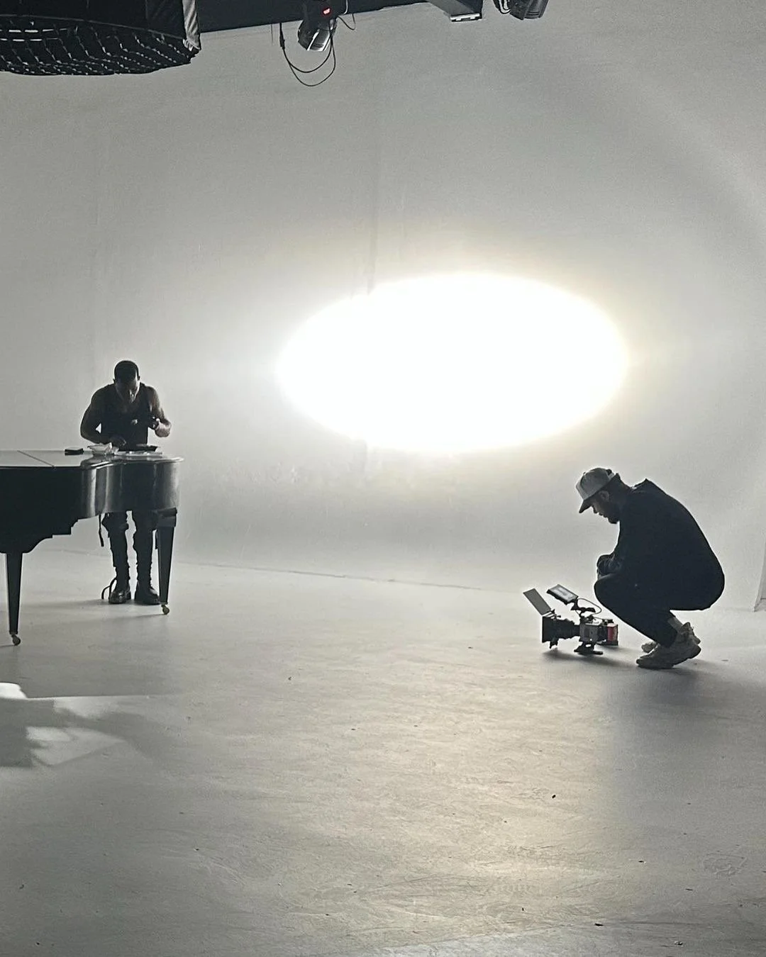 People in a studio with a white backdrop, one person playing a grand piano, another person squatting with camera equipment, and studio lights overhead.
