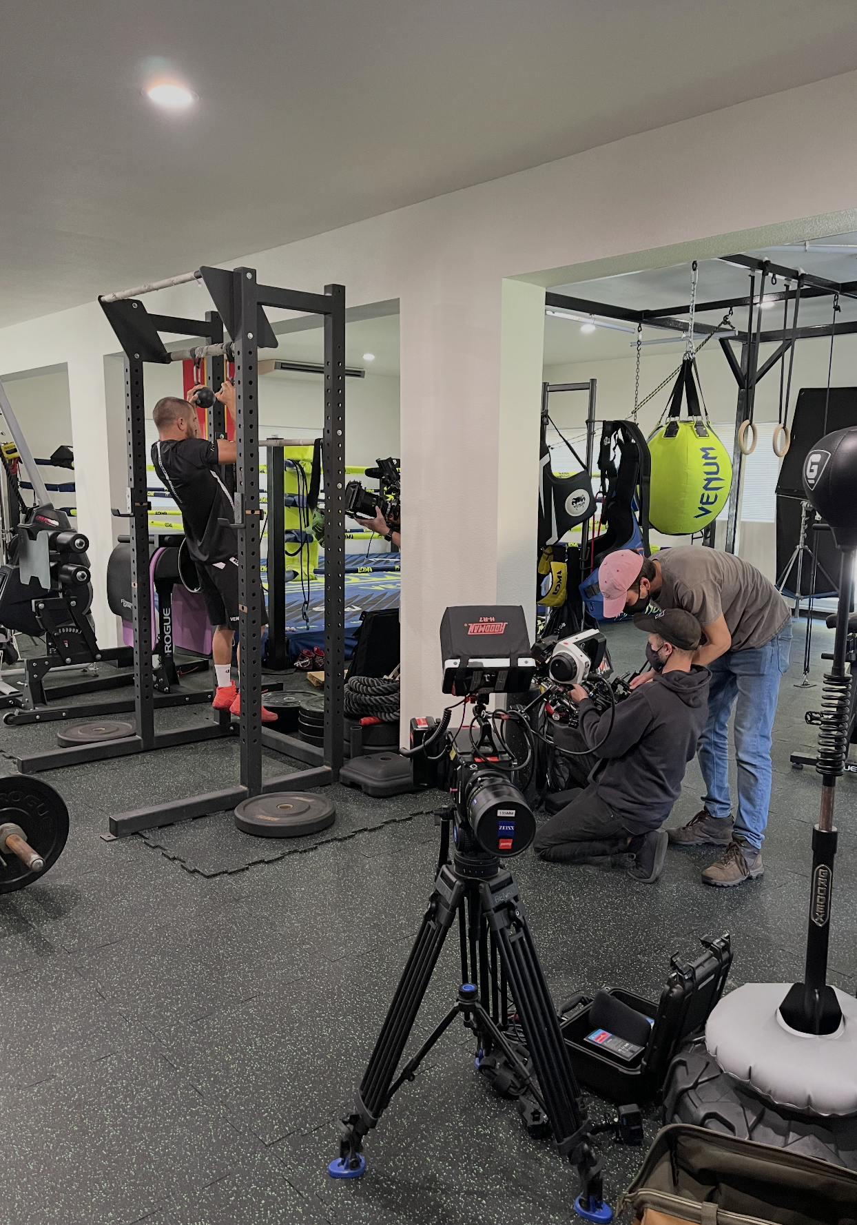 A man working out on a squat rack with weights in a gym while being filmed by a camera crew. Two crew members are behind the camera, one adjusting the camera and the other leaning over.
