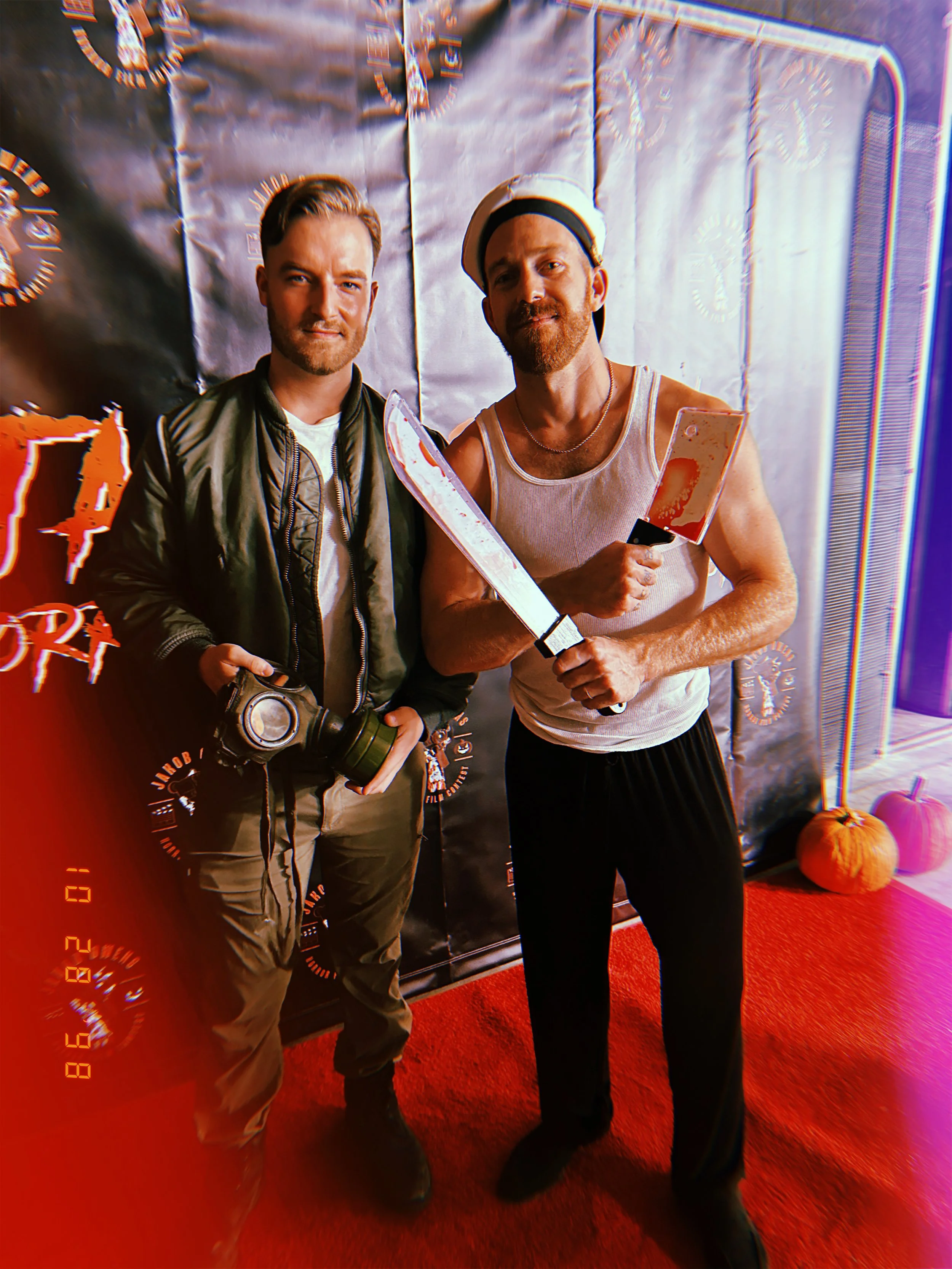 Two men posing for a photo at a Halloween party, one holding a camera and the other a knife and a blood-stained cleaver. They are standing on a red carpet in front of a black backdrop, with pumpkins on the floor to the right.