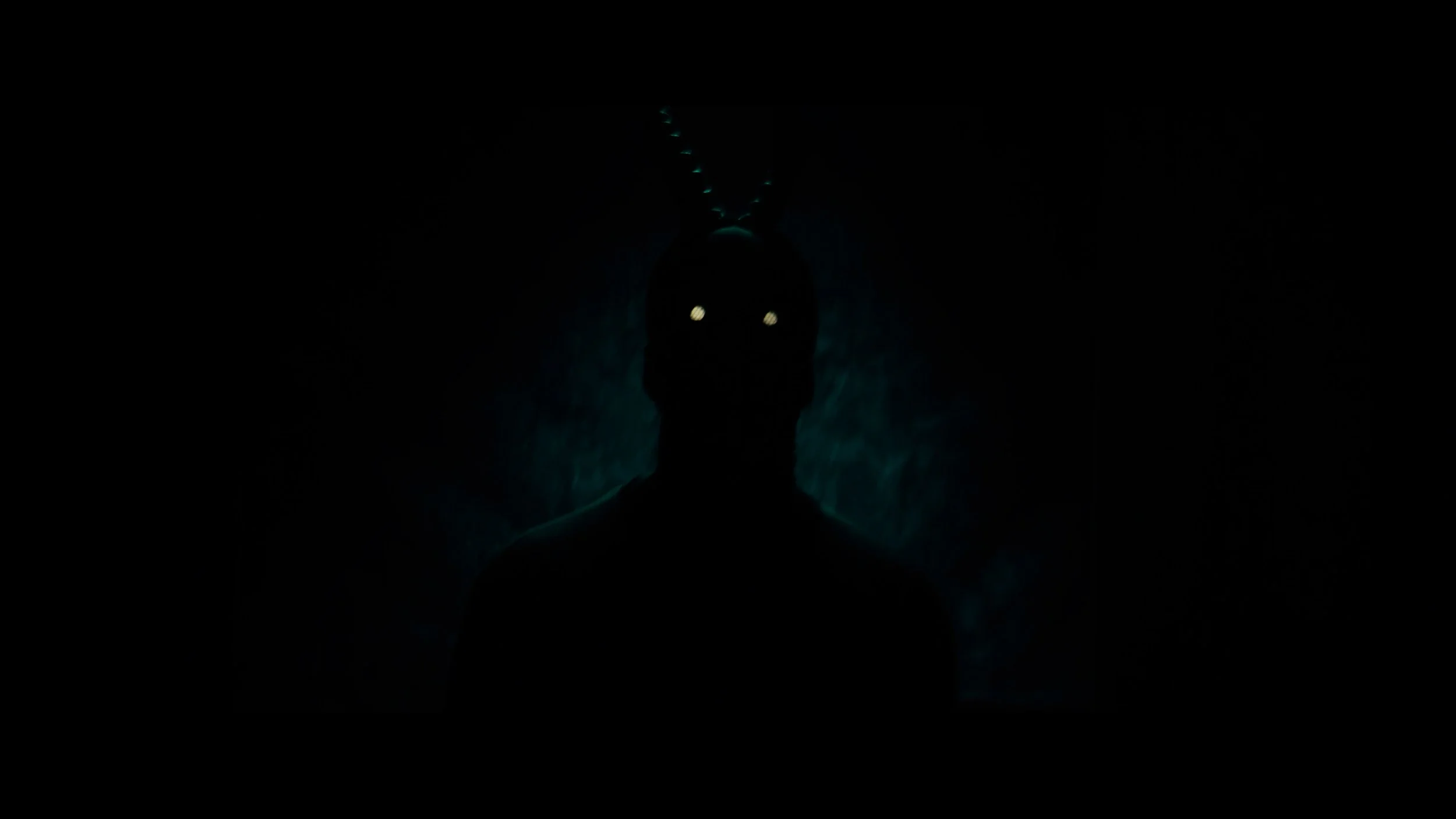 Dark silhouette of a person with glowing eyes, surrounded by an eerie, smoky blue-green glow, and antennae-like protrusions on the head.