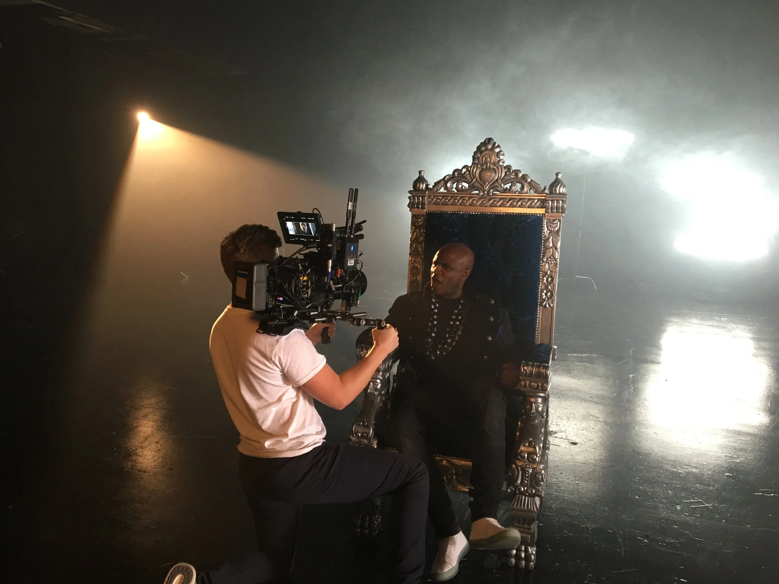 A person operating a camera films a man sitting on an ornate throne in a dark, smoky studio