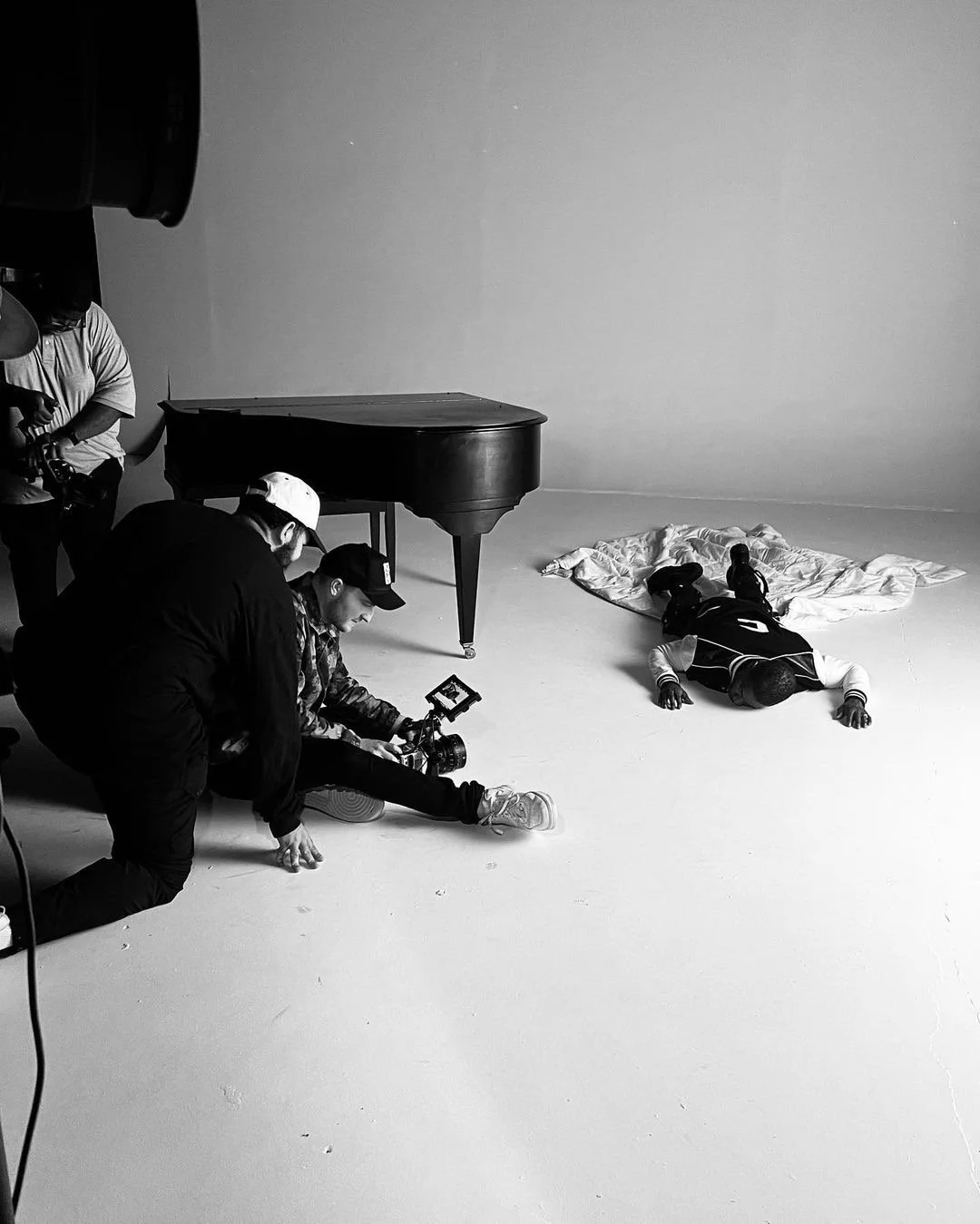 Black and white photo of a person lying on the floor with eyes closed, head turned to the side, covered with a white blanket, and another person assisting or filming, with a camera, in a studio setting with a piano and plain background.