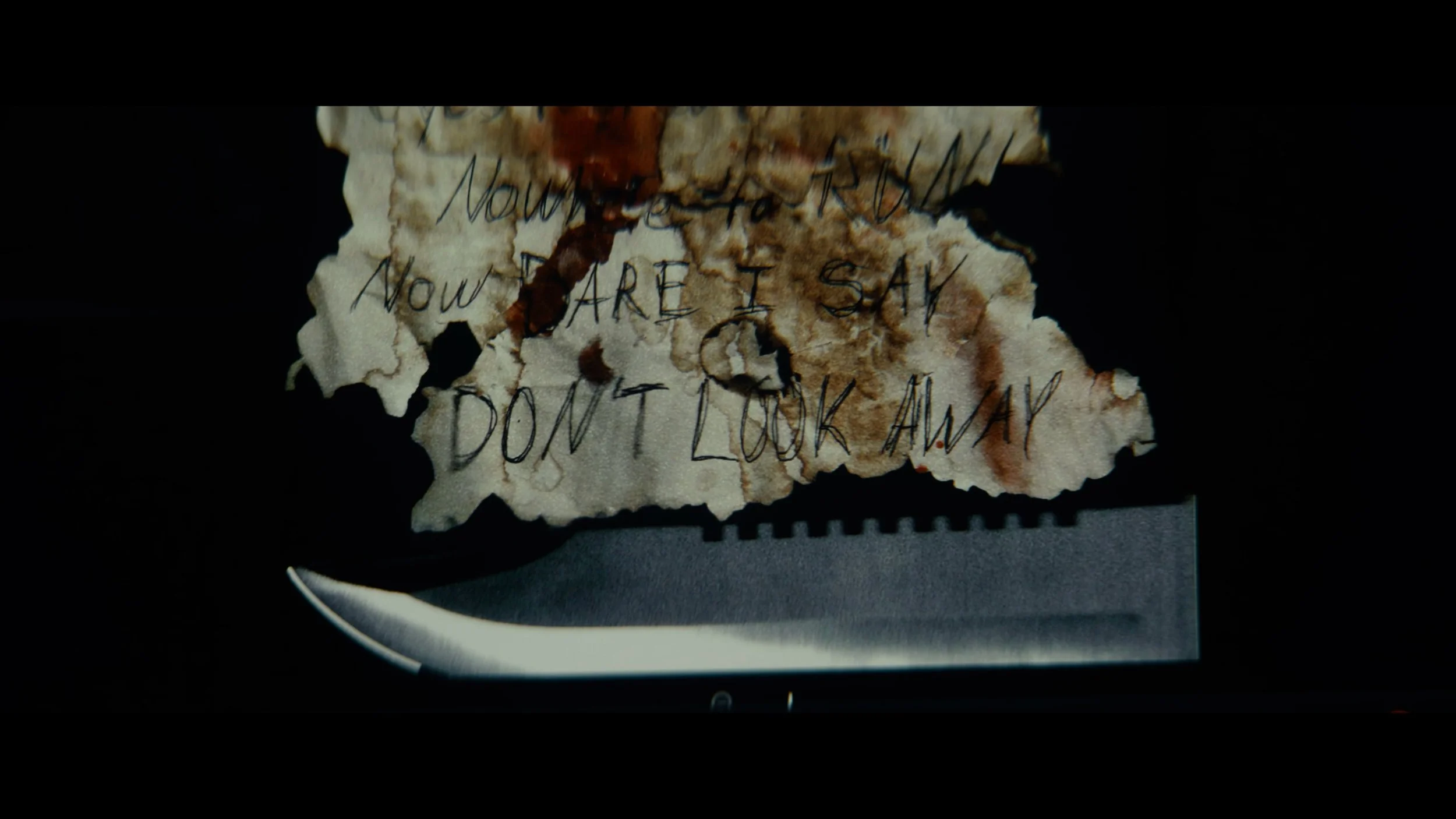 Torn, burnt piece of paper with handwritten message that says, 'Now you to know, now I are I say, don't look away.' on a dark background with a knife at the bottom.