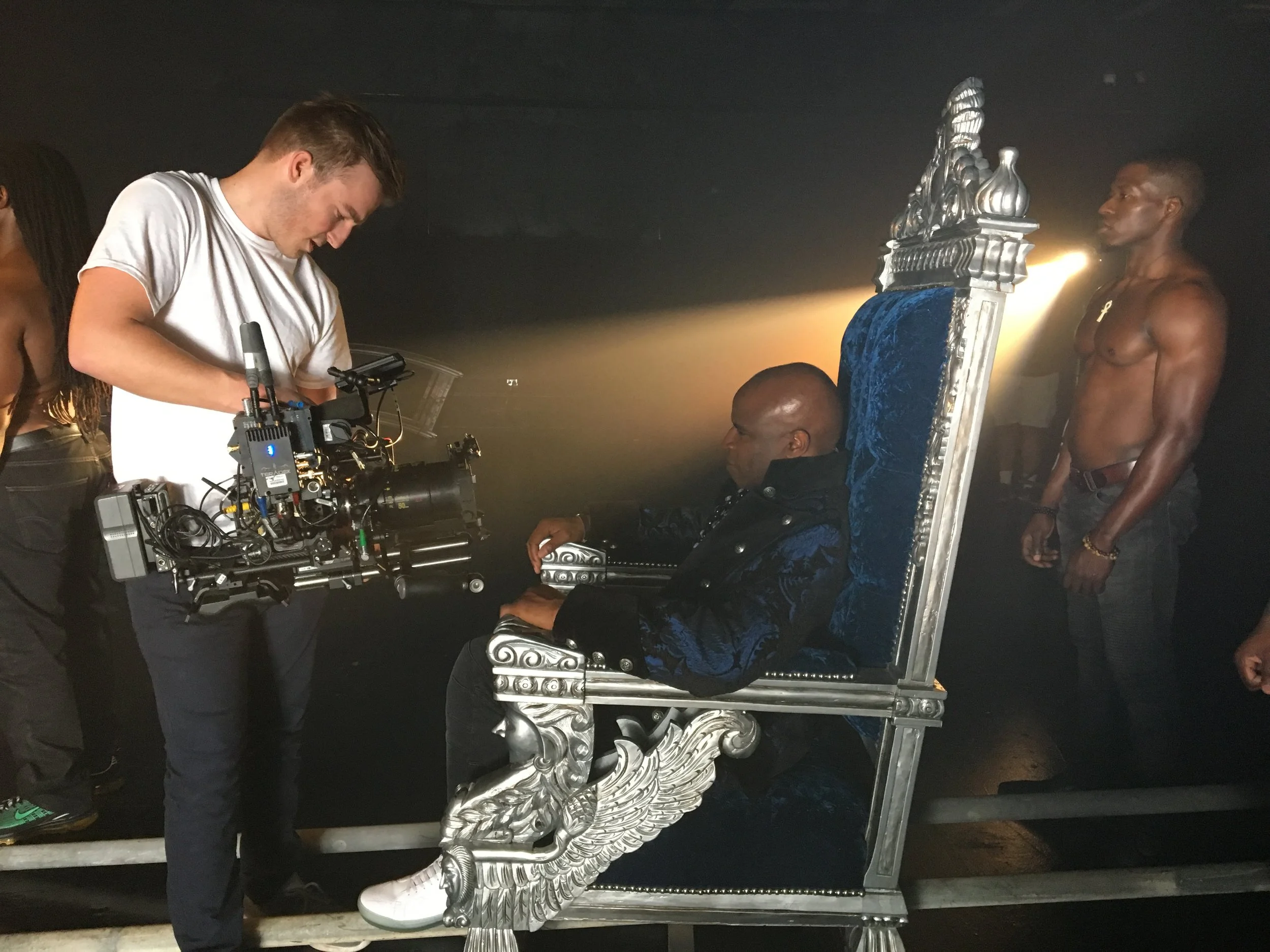 Filming scene with a man seated on an ornate throne-like chair, crew member with a camera standing next to him, and another shirtless man standing nearby.