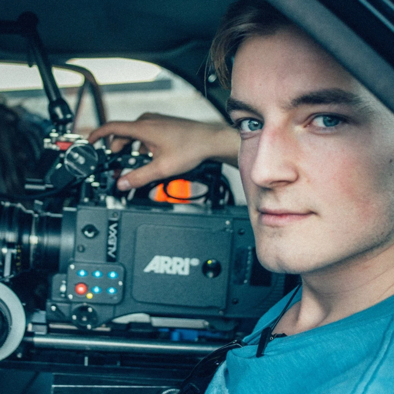 Close-up of a young man with blue eyes and brown hair operating a professional ARRI camera, inside a vehicle or a confined space.