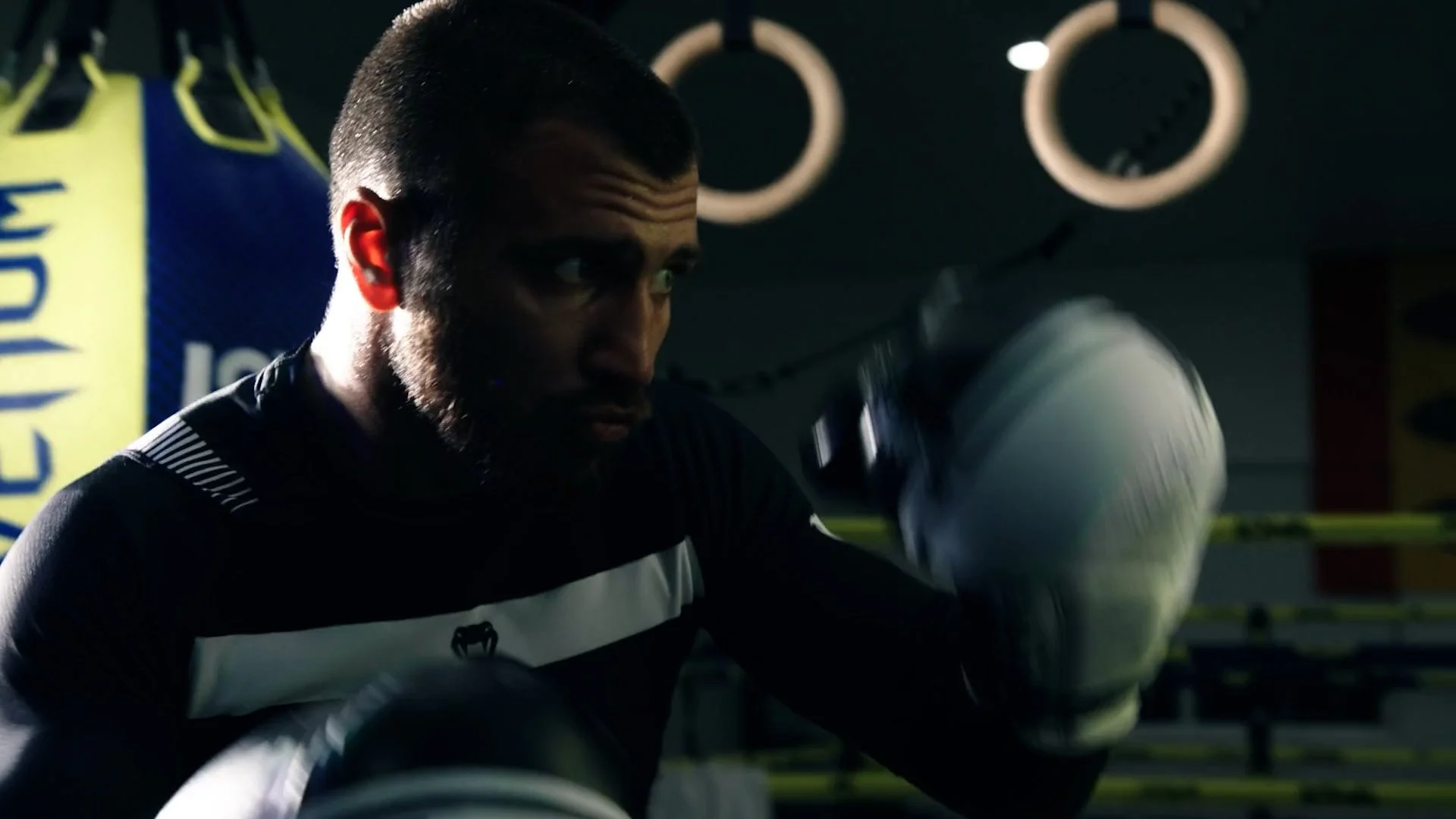 A man wearing boxing gloves is training in a gym, with a boxing ring and a yellow banner in the background.