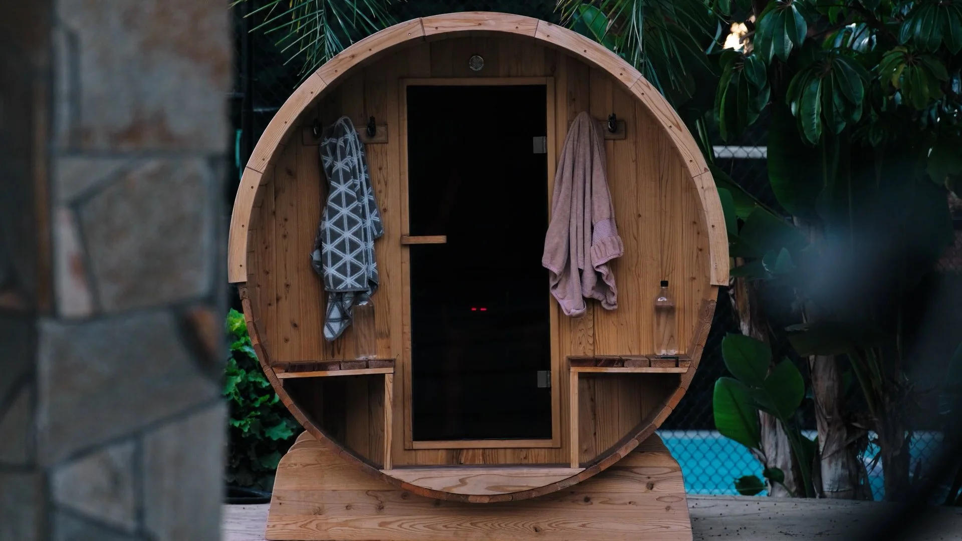 Wooden barrel-shaped outdoor sauna with hanging towels and bottles, surrounded by greenery.