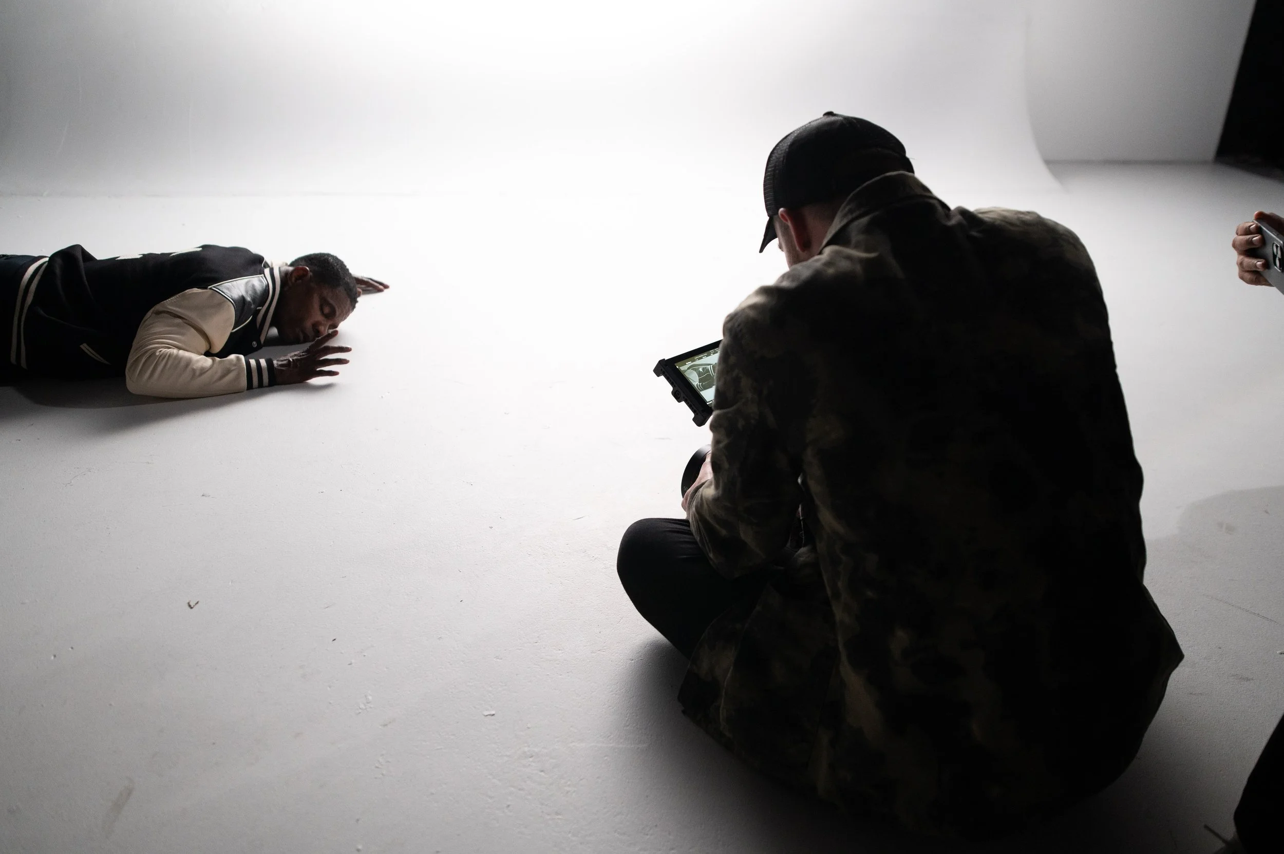 Photograph of a photoshoot setup with a male model lying on a white studio floor, and a photographer taking pictures while another person holds a phone nearby.
