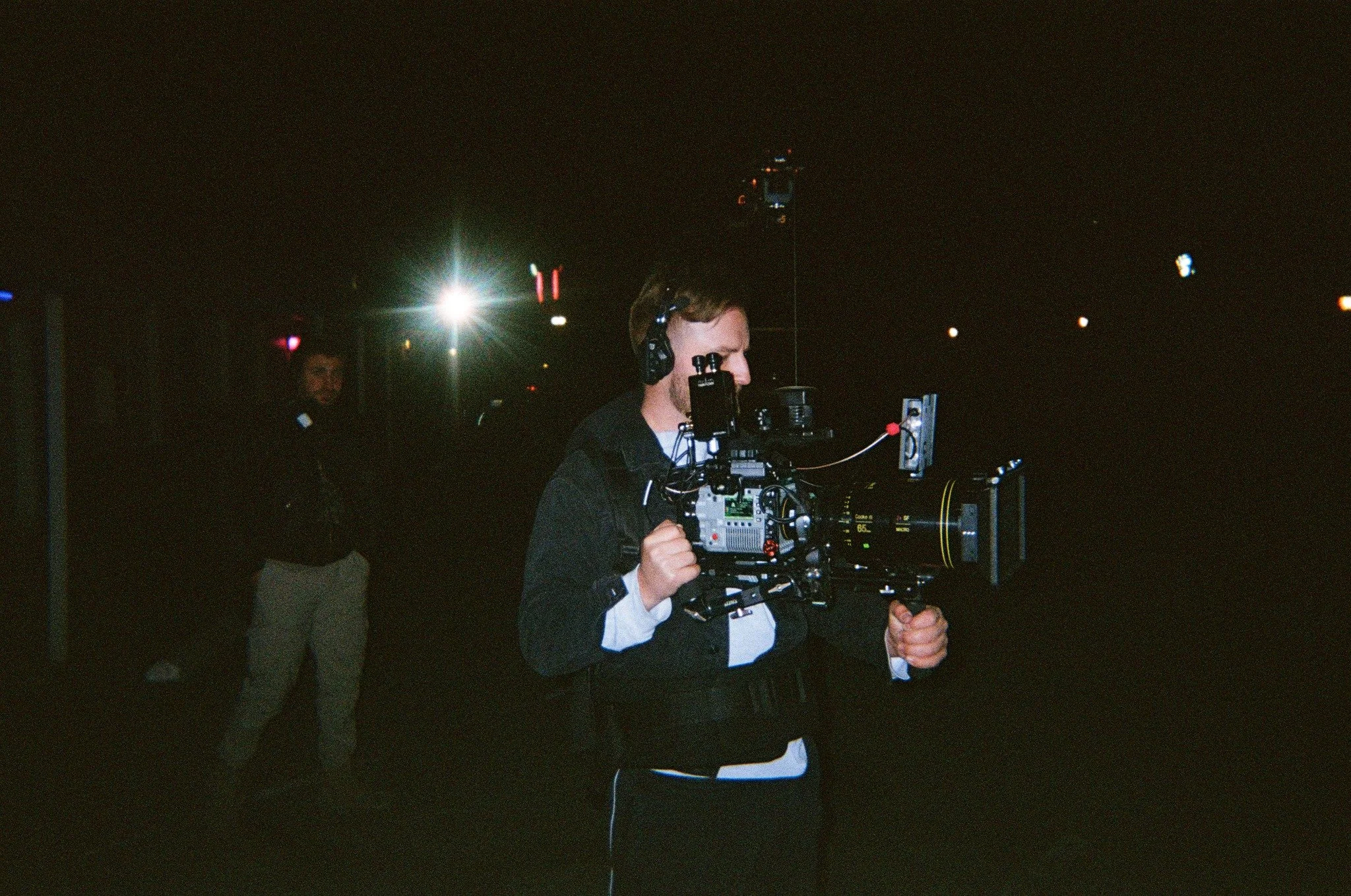 A man wearing headphones operating a professional video camera at night. Another person is in the background with a bright light behind them.