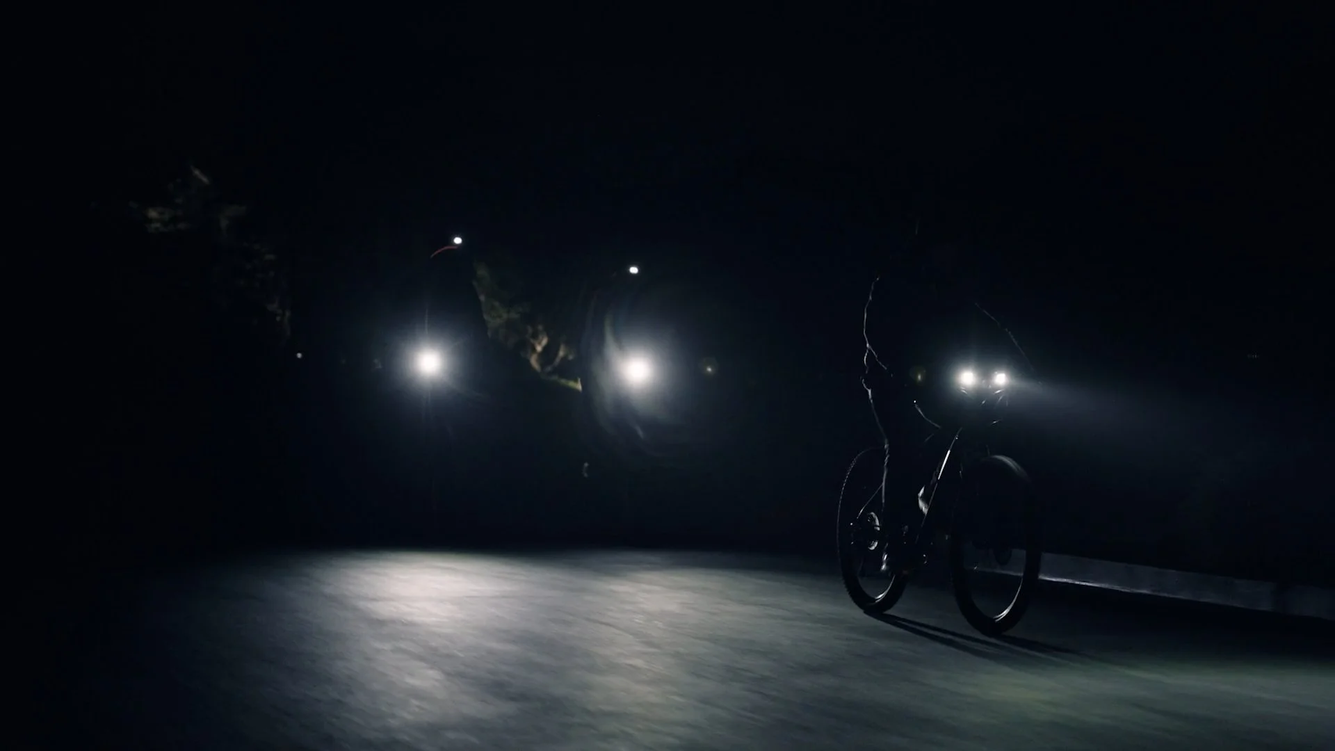A cyclist riding a bike at night with headlights on, illuminating the dark road and trees in the background.