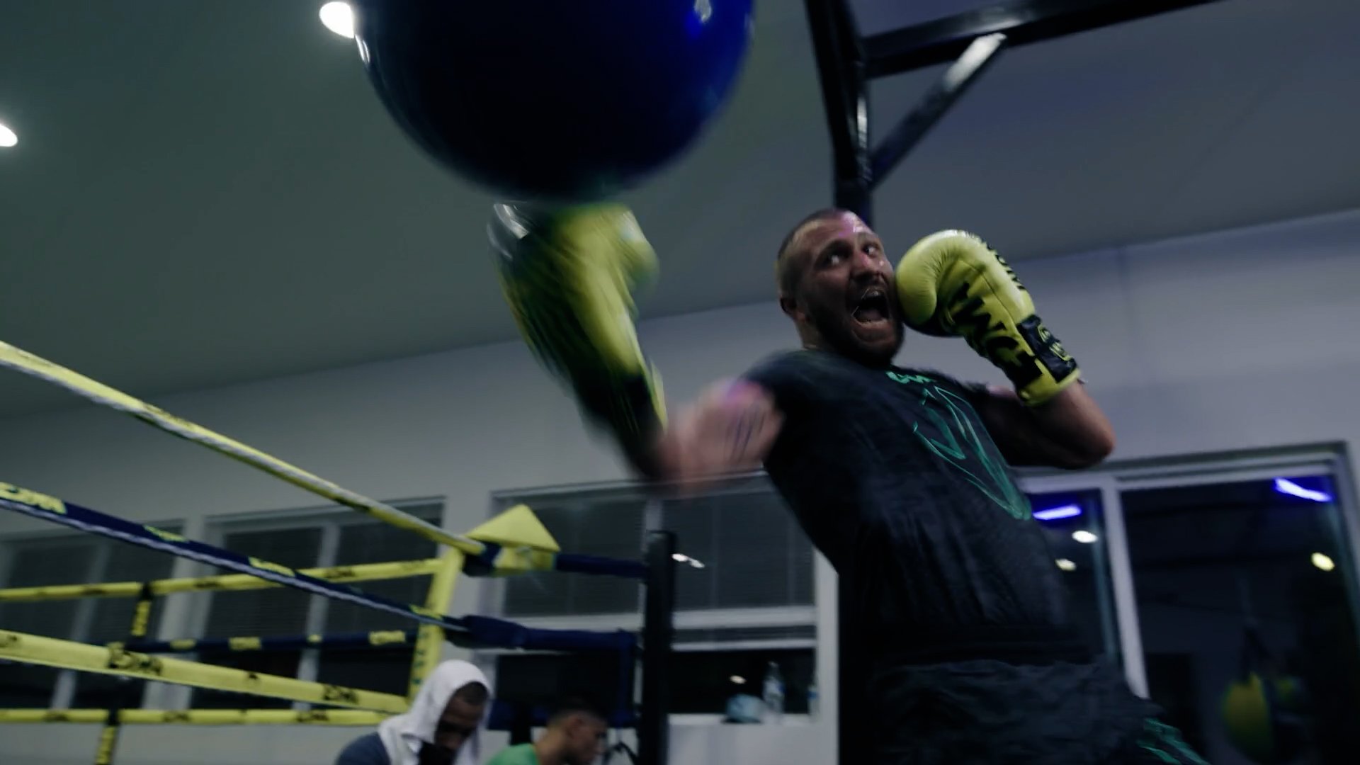 A man wearing black athletic clothing and yellow boxing gloves is punching a hanging heavy bag in a gym. He looks intense and focused.
