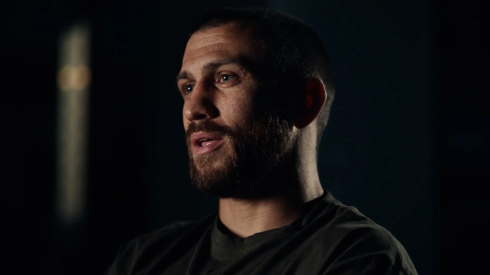 Close-up of a man with a beard speaking, against a dark background.