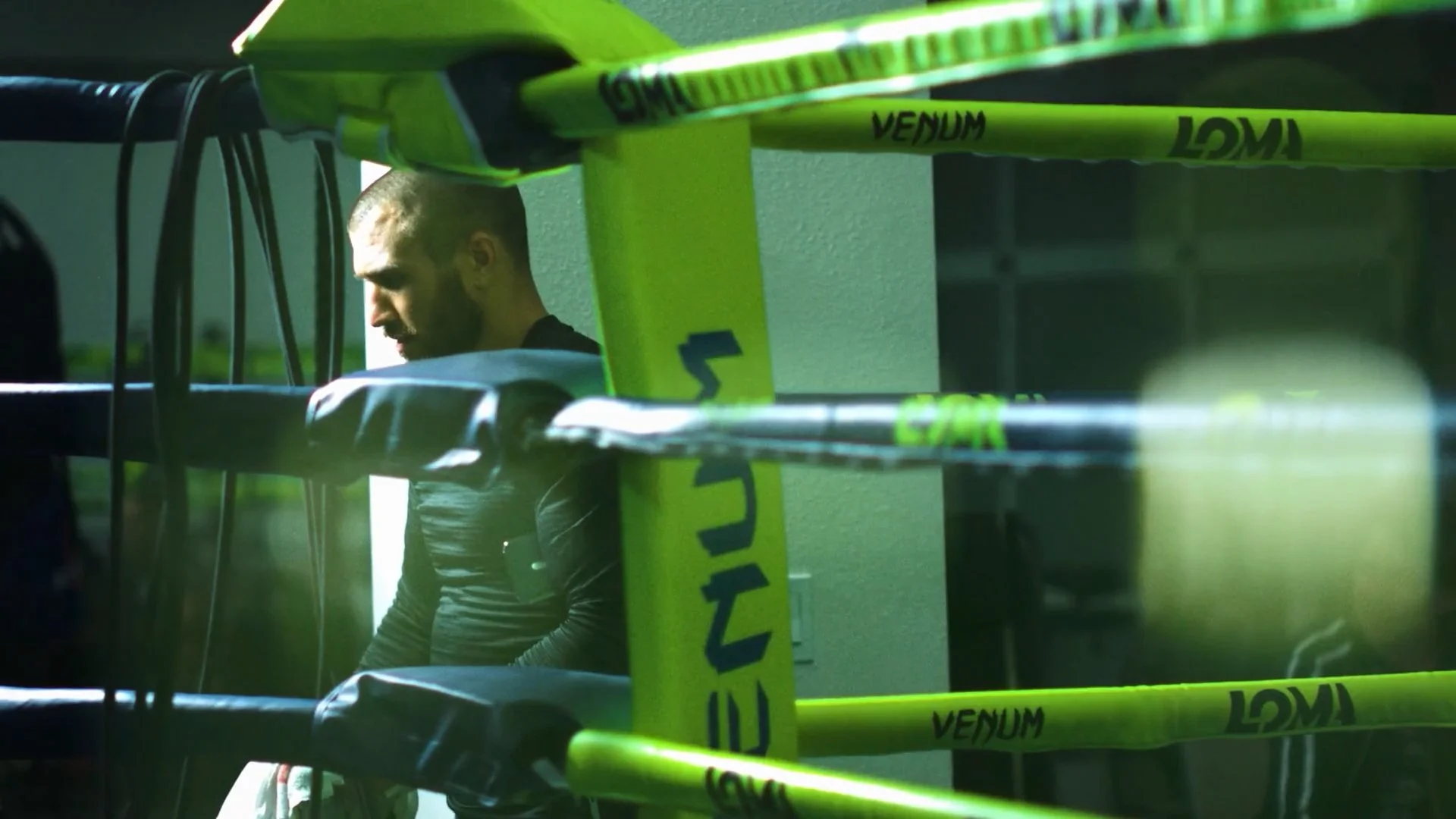 A male athlete in boxing gloves and athletic gear sitting in a boxing ring, with the focus on the side of the ring and yellow branding tape marked with 'VENUM' and 'LOMA'.