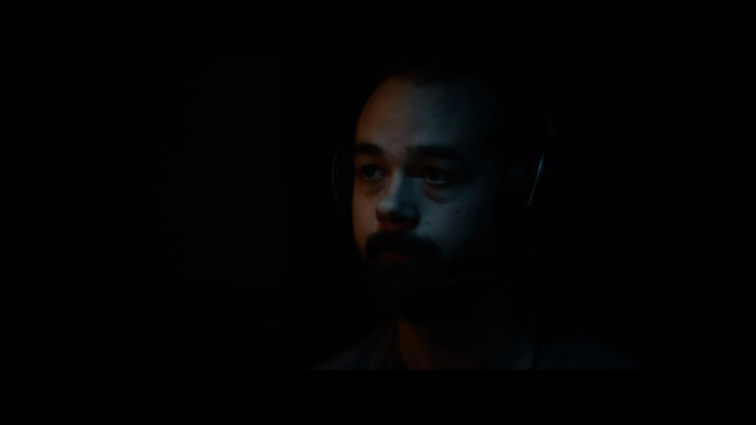 A man with facial hair and wearing headphones in a dark room, illuminated by a subtle light on one side of his face.