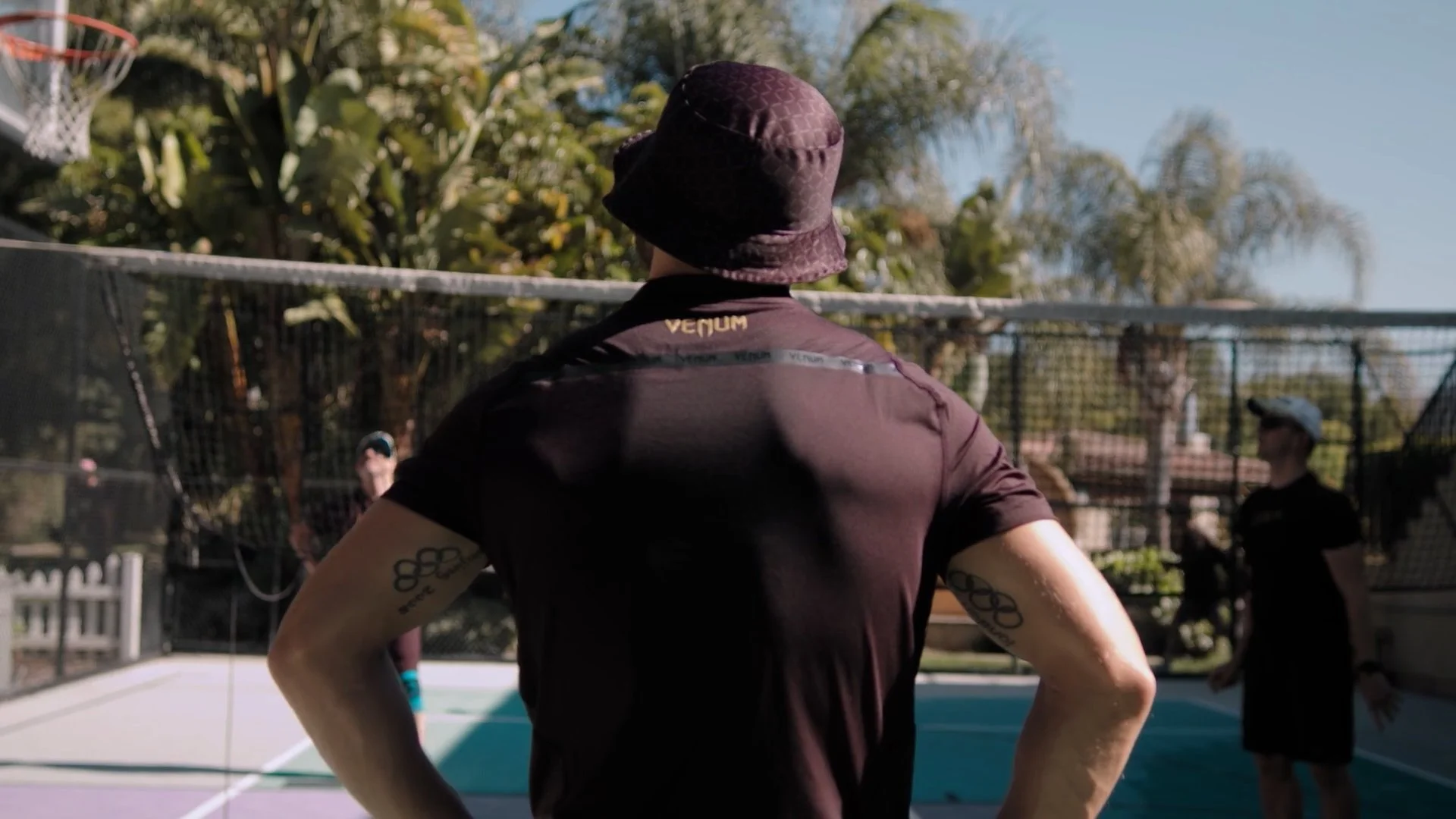 A person wearing a black shirt and purple bucket hat stands on a tennis or pickleball court, facing the net, with their back to the camera. Several other people are visible in the background, and it is a sunny day with trees and a blue sky.