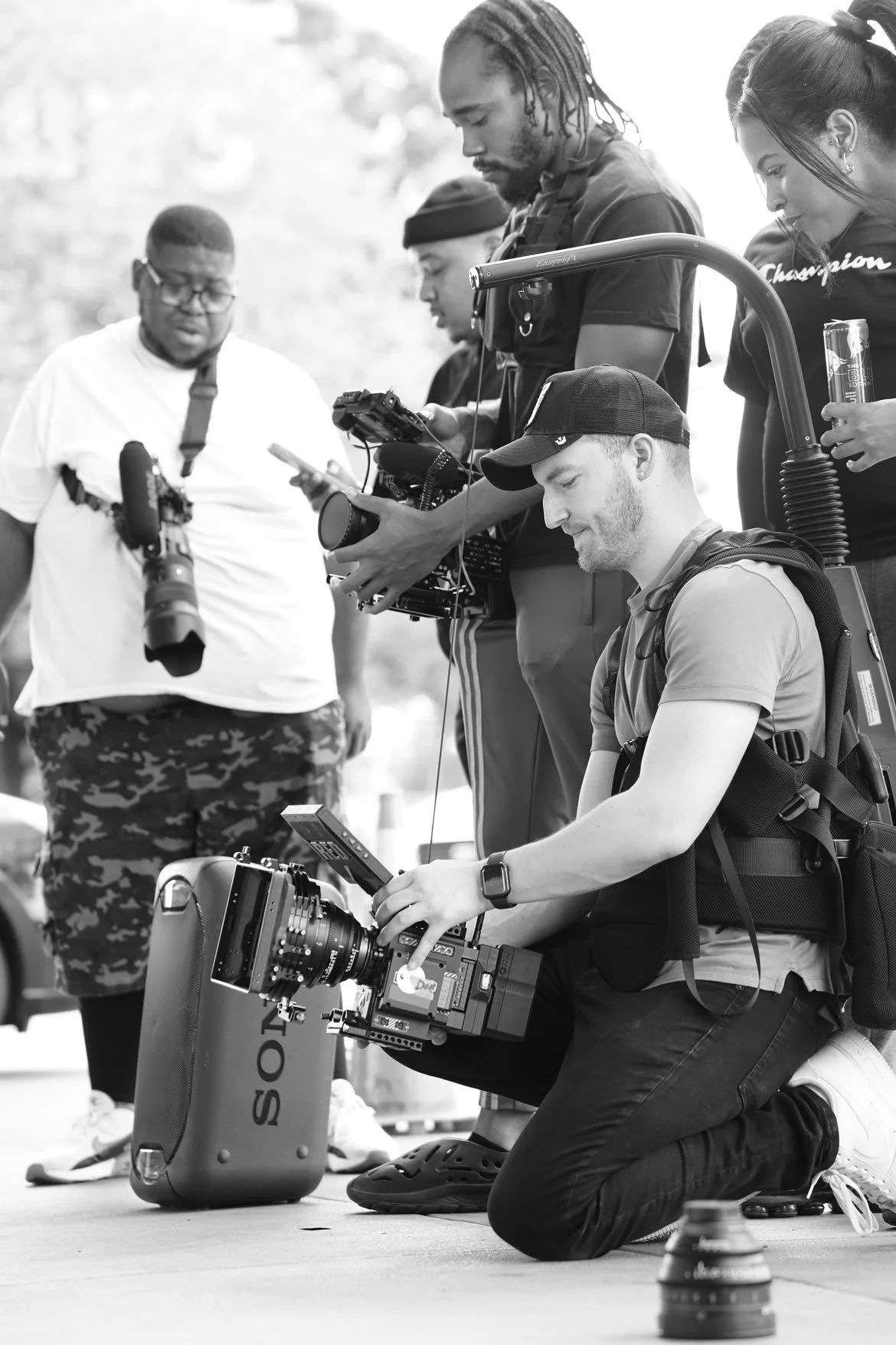 Group of people, including a man kneeling with professional camera equipment, filming or photographing outdoors during the daytime.