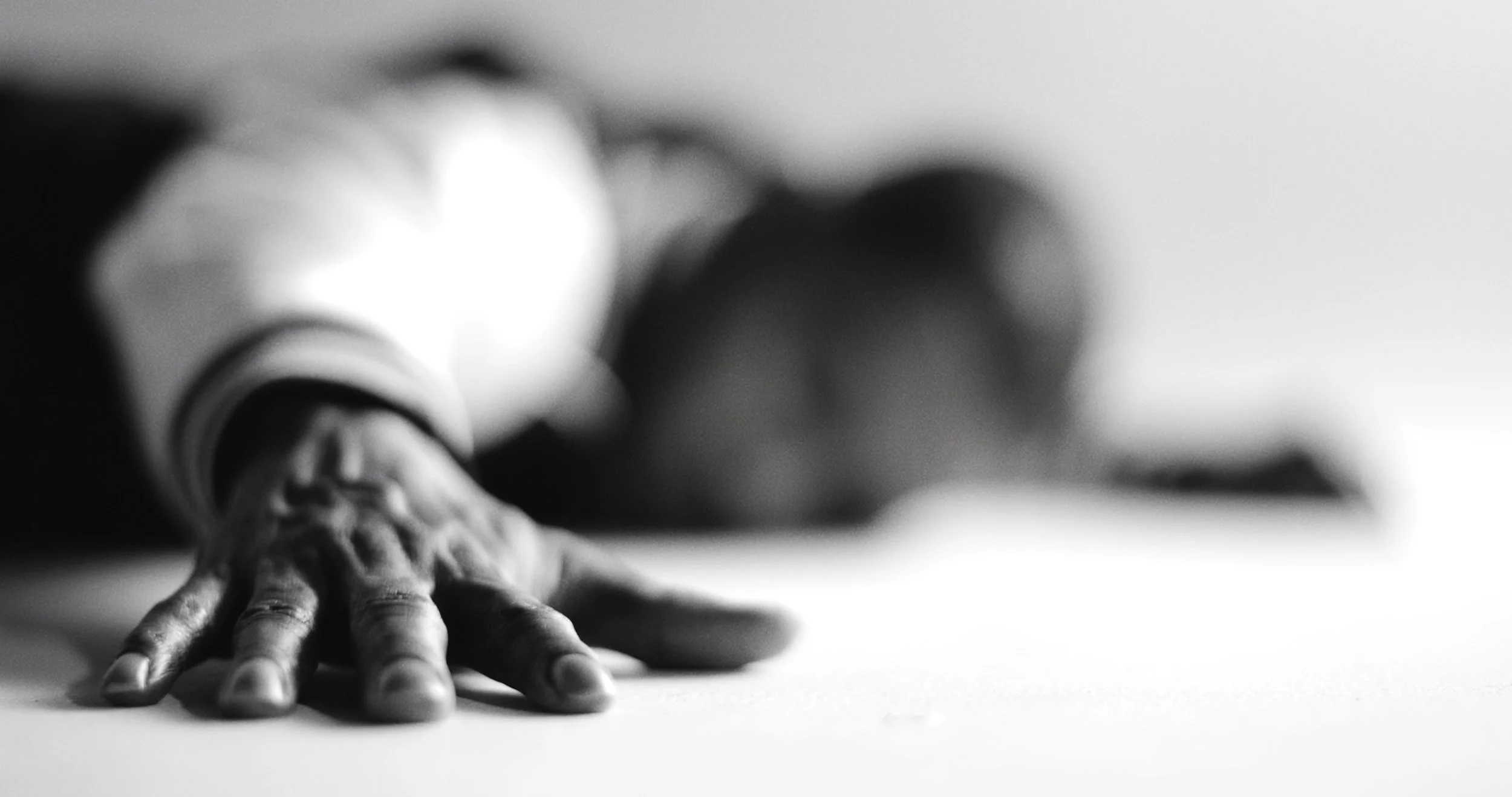 Black and white photograph of a person lying face down with their hand stretched out toward the camera, fingers splayed.
