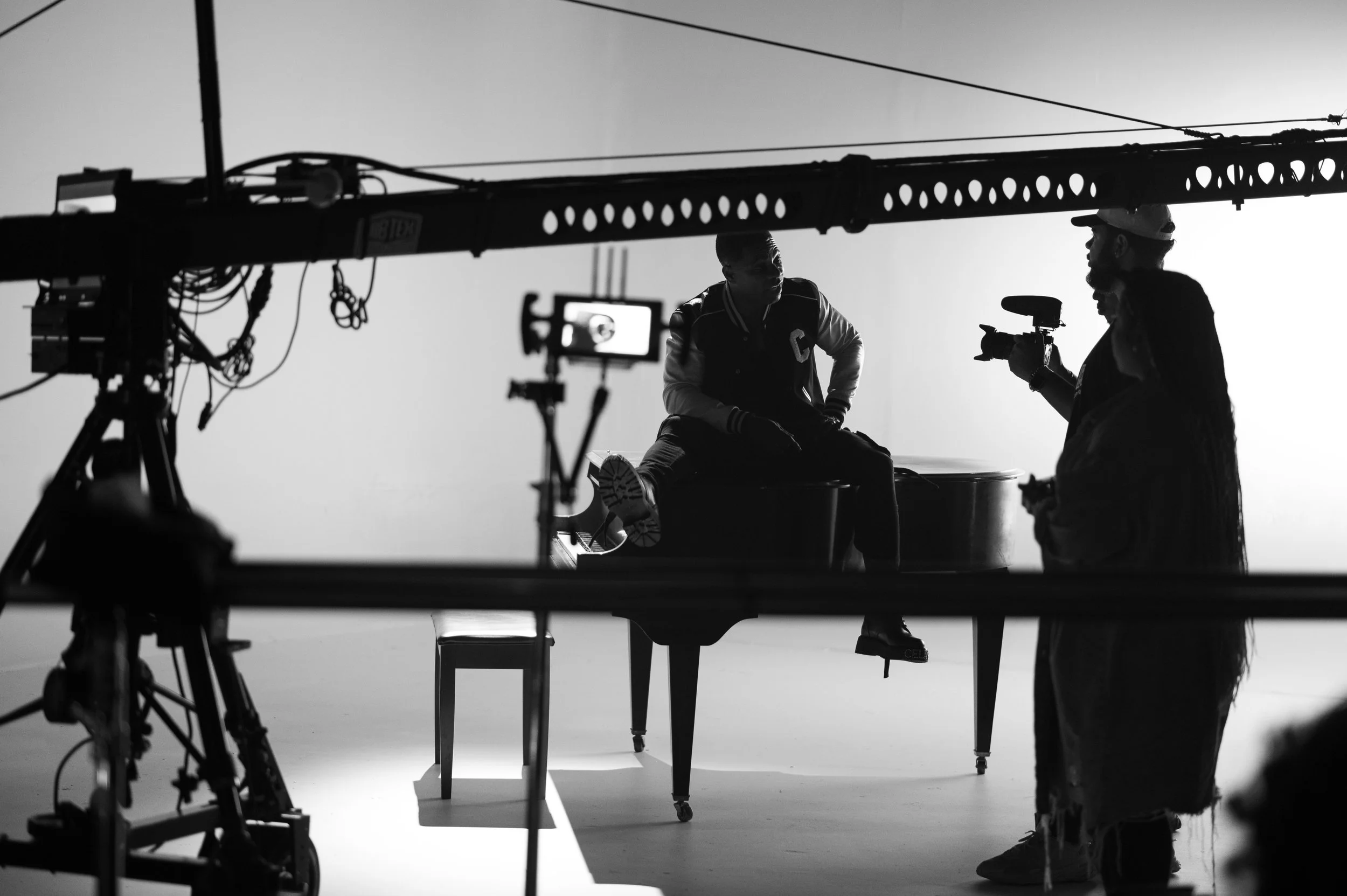 Black and white photo of a film set with three people. One person is sitting on a piano, another is holding a video camera, and the third person is standing, all engaged in conversation. Film equipment and lighting are visible.