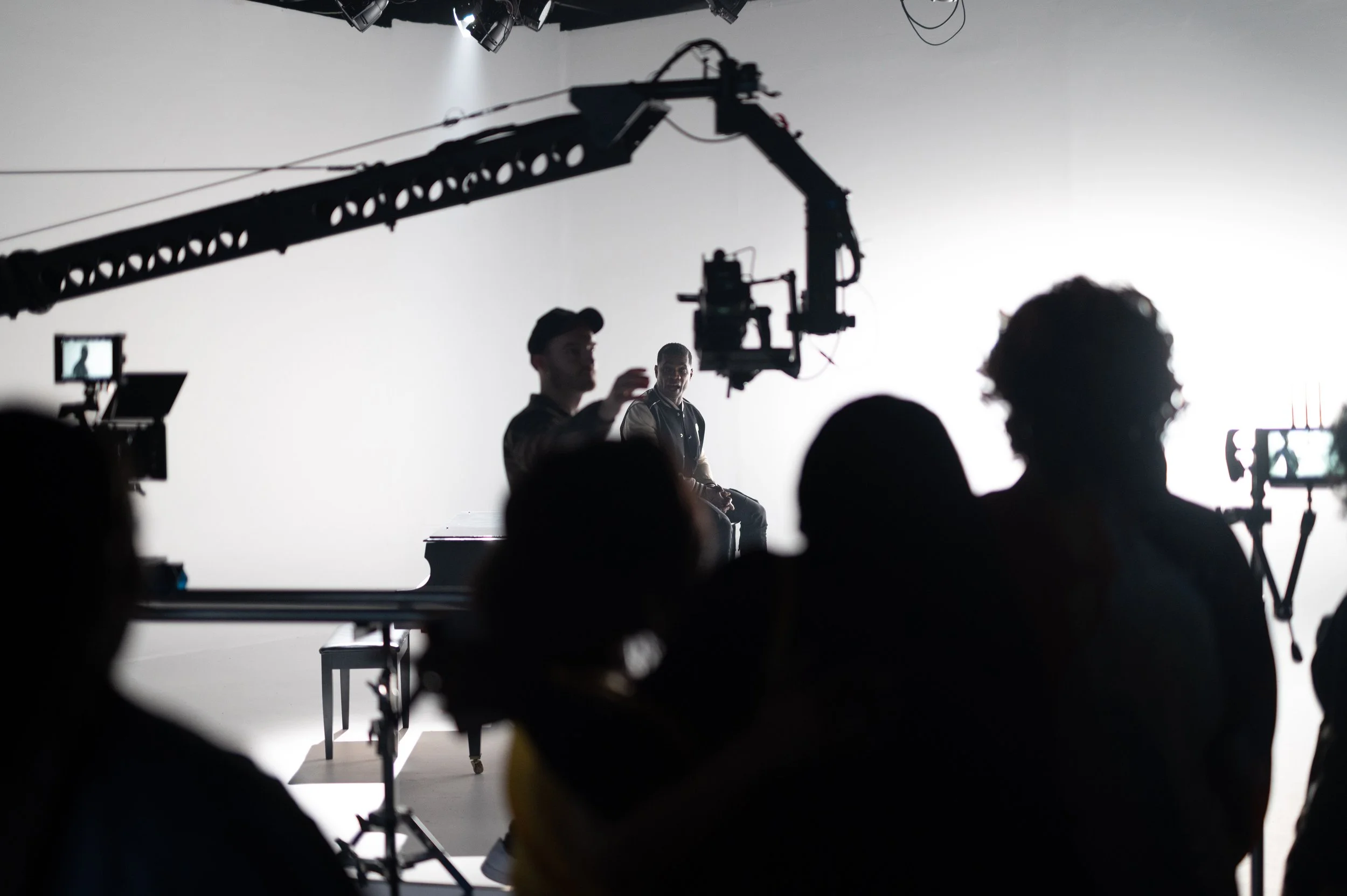 Filmmaking scene with two actors on set, cameras, and crew, against a white backdrop.