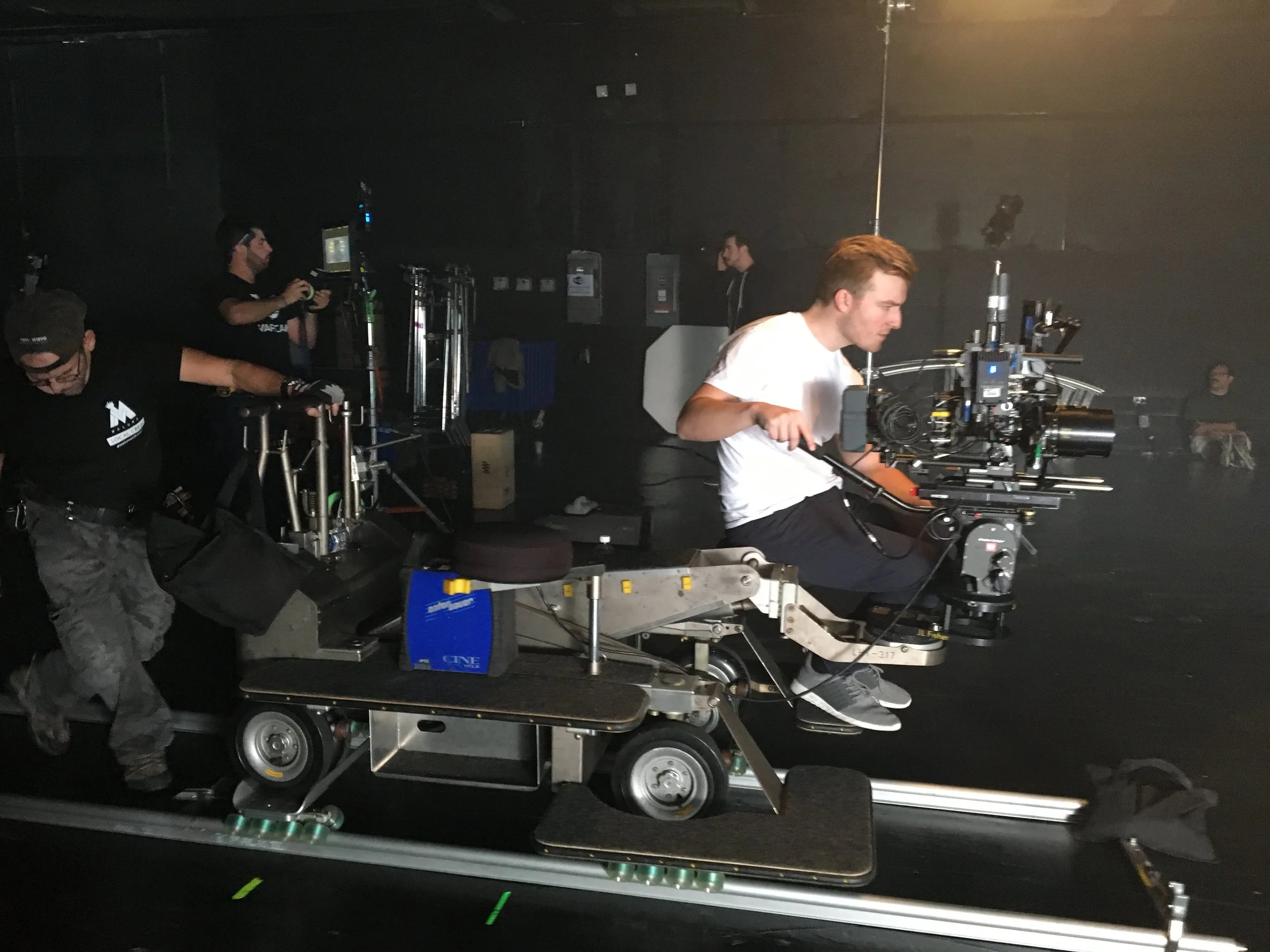 A person operating a large camera on a dolly during a film shoot in a dark studio.