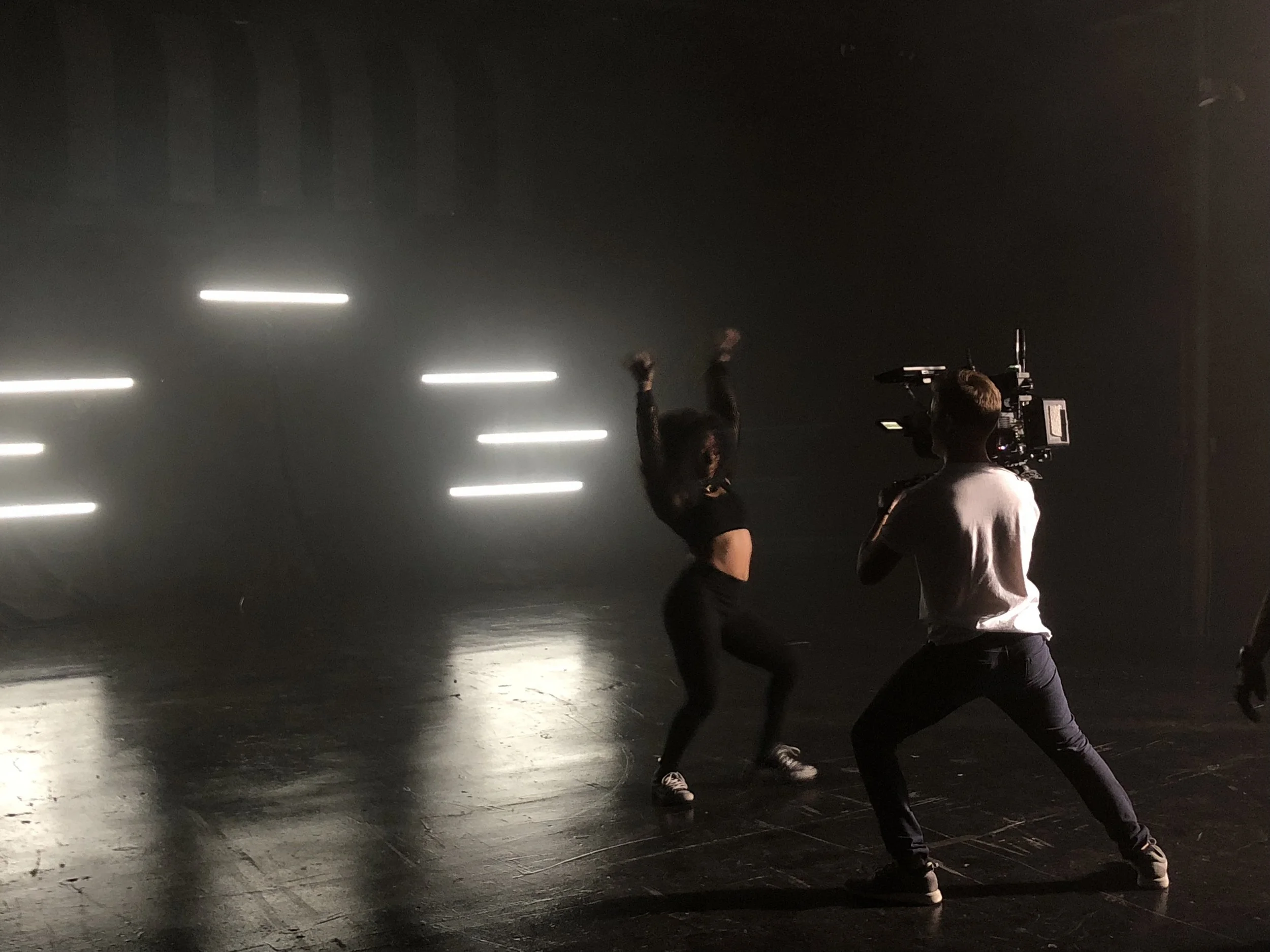A person with a camera filming a dancer in a dark, foggy studio with bright horizontal lights in the background.