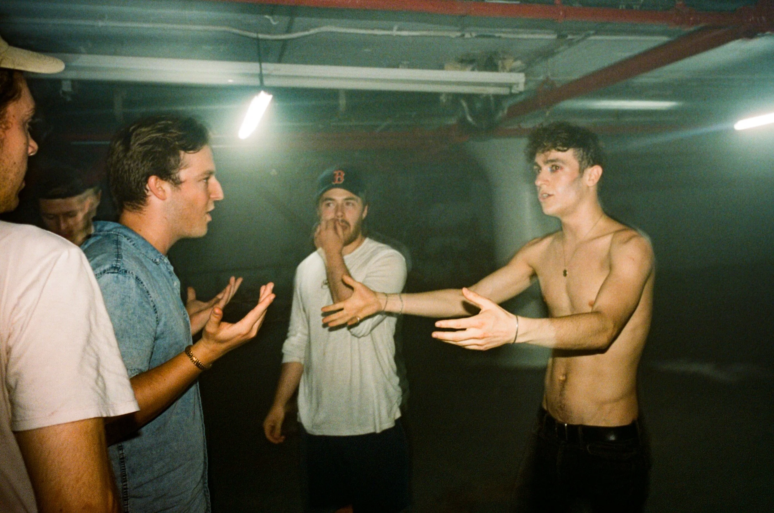 A group of five young men are having a conversation in a dimly lit indoor space, with one shirtless man gesturing as he speaks, and others listening and reacting.
